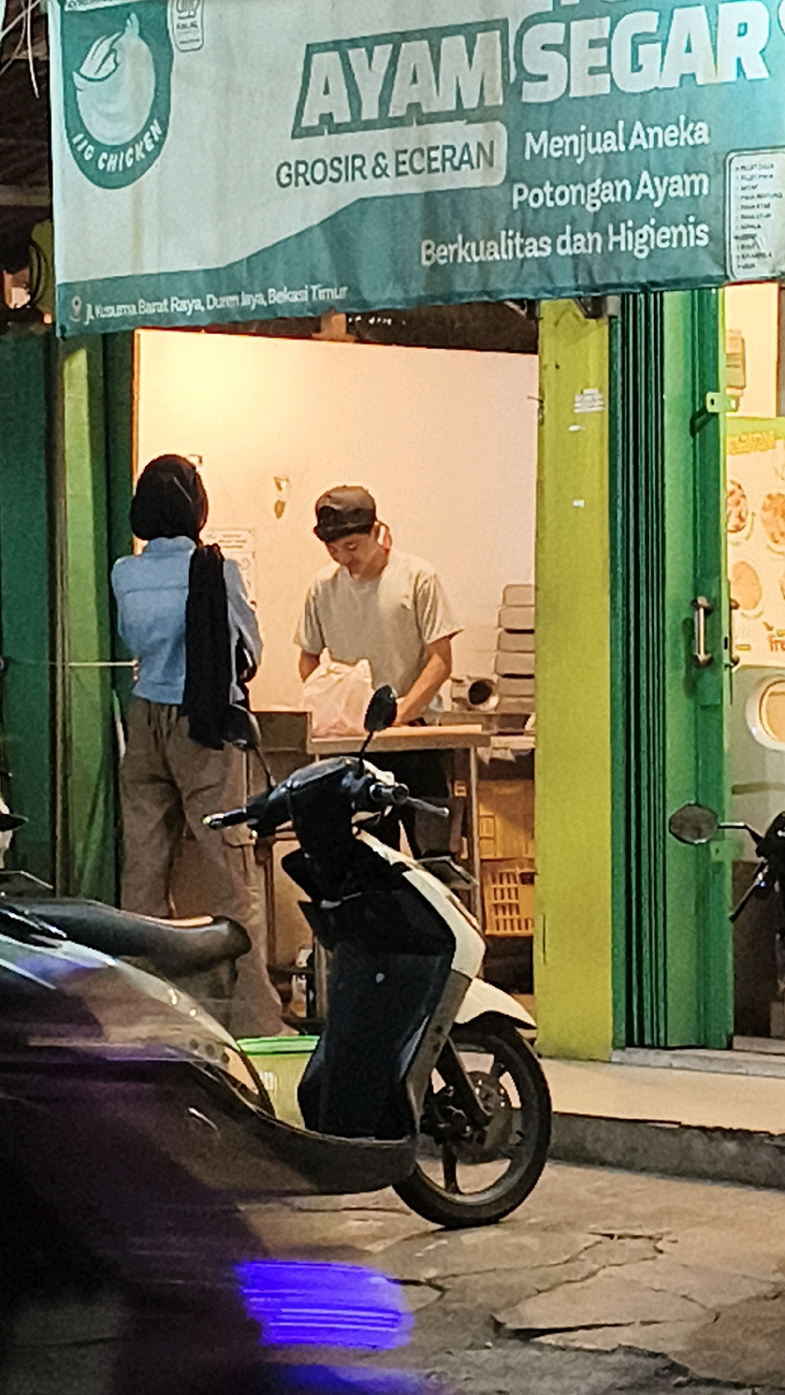 The image captures an outdoor scene at night in Bekasi, Indonesia, likely in front of a food stall or small business. A banner above the entrance reads "AYAM SEGAR" (Fresh Chicken) and lists "GROSIR & ECERAN" (Wholesale & Retail), along with "Menjual Aneka Potongan Ayam Berkualitas dan Higienis" (Selling Various Quality and Hygienic Chicken Cuts). Below the banner, an address is visible: "Jl. Kusuma Barat Raya, Duren Jaya, Bekasi Timur."
In the foreground, a dark-colored motorcycle is parked, partially obscuring a purple light reflection on the cracked pavement. Behind the motorcycle, a person wearing a hijab and a light blue denim jacket stands with their back to the camera, facing the interior of the establishment. A dark scarf is draped over their right shoulder.
Inside the establishment, a person wearing a light-colored t-shirt and a dark baseball cap is visible behind a counter. They are looking down and appear to be handling a plastic bag filled with items. Stacks of white boxes are visible behind them, and to their right, a green accordion-style door is partially open, revealing the interior. The overall environment suggests a humble, street-level business.