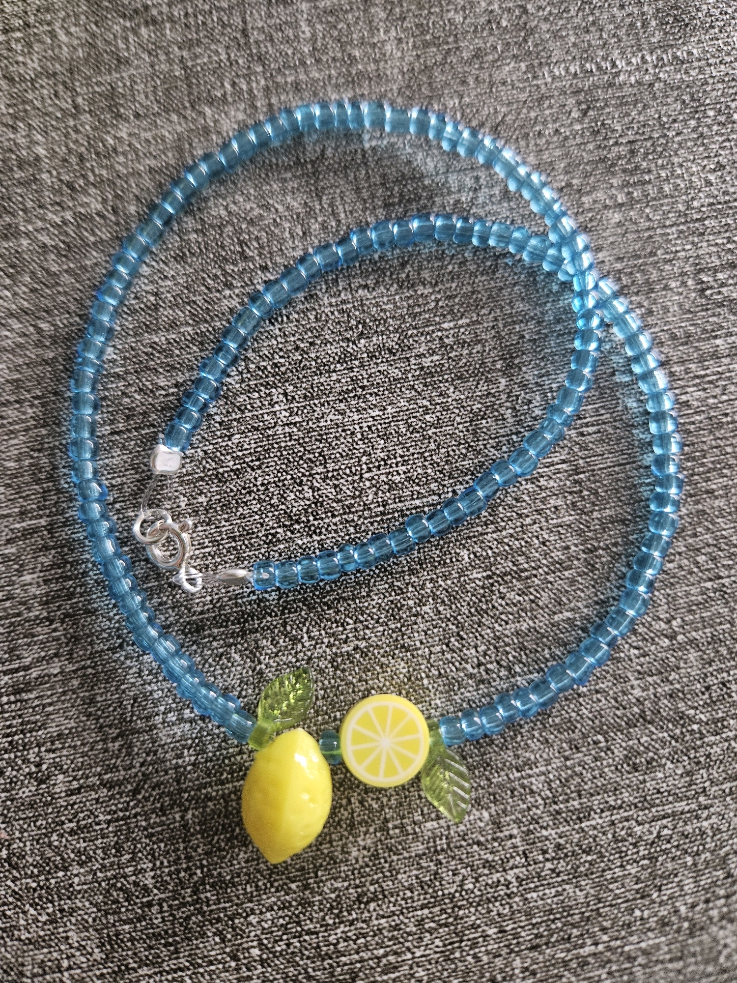 A blue beaded necklace with a lemon charm and leaf charms lies on a gray, textured surface. The necklace is open and forms a loose loop. The lemon charm is yellow and shaped like a whole lemon, with a smaller yellow lemon slice charm next to it. Two green leaf charms are attached to the necklace, one on either side of the lemon charms. The necklace has a silver clasp. The background appears to be a fabric or textured surface. No specific location in Burgas, Bulgaria can be determined from the image.