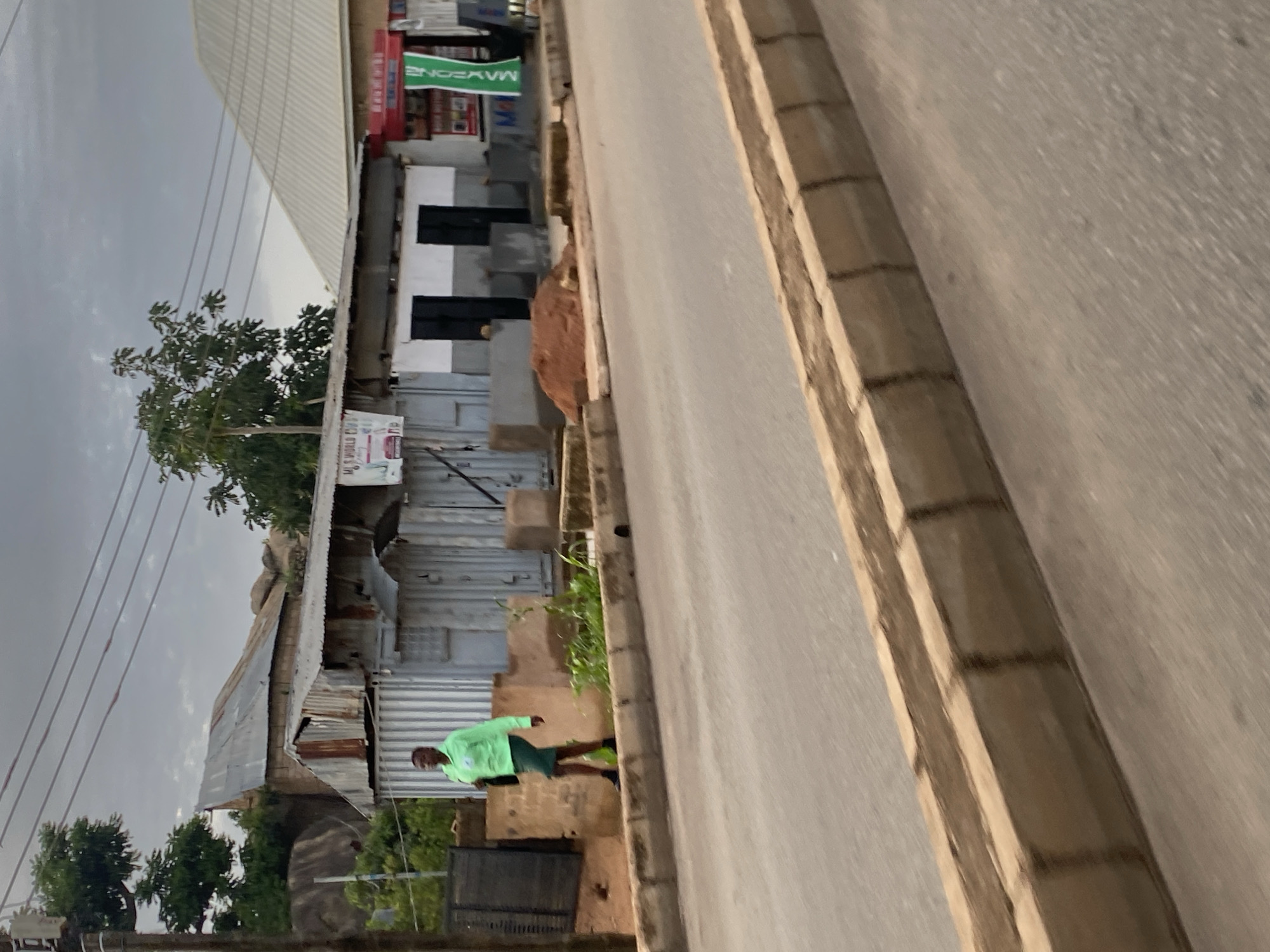 The image shows a street scene in Barakin Lerawi, Nigeria. A person wearing a bright green jacket and dark shorts is walking on the sidewalk next to a road. The sidewalk is lined with a concrete curb. In the background, there are several buildings, some with corrugated metal roofs. One building has a sign that reads "MJ'S WORLD." Another building has a green banner with the word "MAYZONE" on it. There are also some trees and large rocks visible. The sky is overcast.