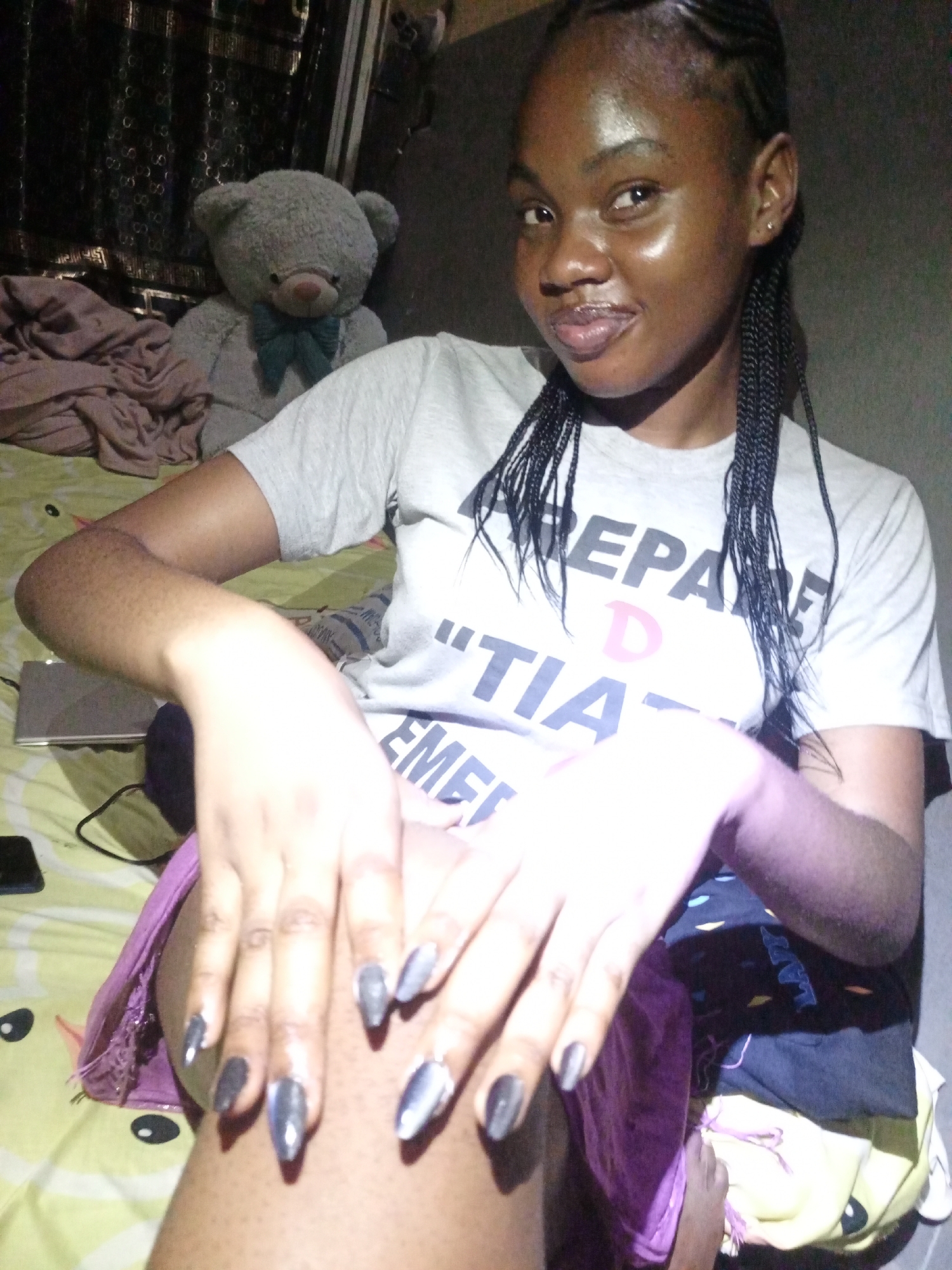 A young woman is posing for a selfie in a room in Keffi, Nigeria. She is wearing a grey t-shirt with black text, and her hair is styled in braids. She has long, pointed, metallic-colored nails. Her hands are positioned in front of her, displaying the nails. Behind her is a bed with patterned bedding, a grey teddy bear, and a curtained window. A laptop and a mobile phone are visible on the bed beside her. The lighting in the room is dim.