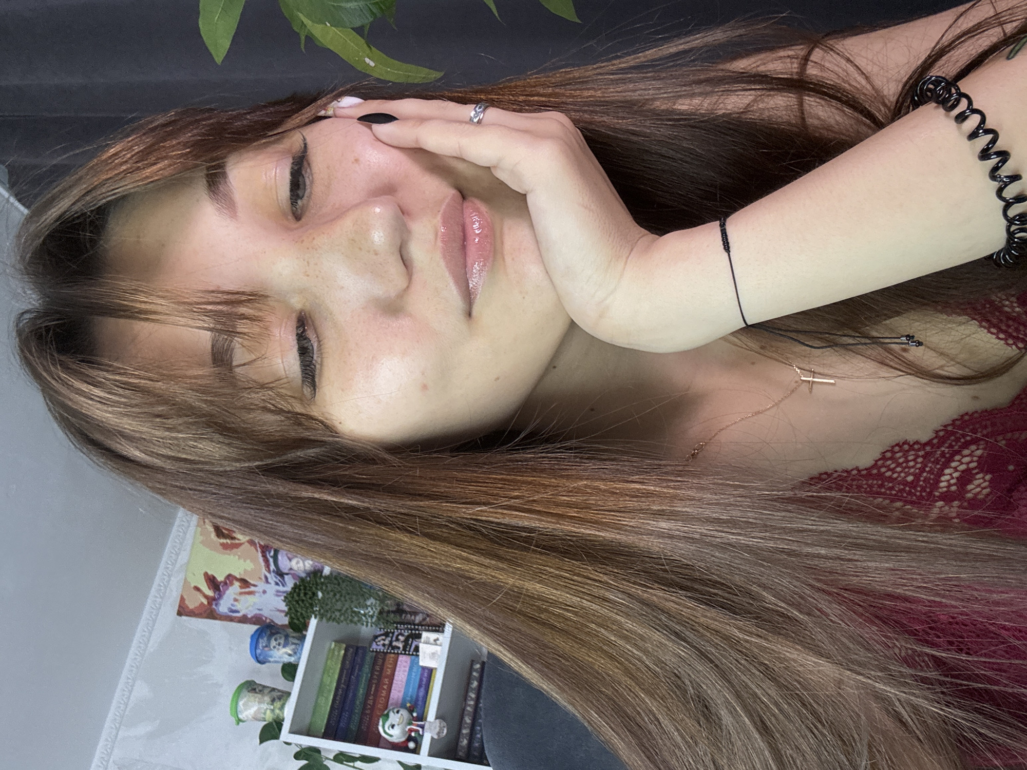 The image is a medium close-up selfie of a young woman with long, brown hair with auburn highlights, and bangs. She is resting her chin on her right hand, which has black nail polish and a silver ring on her index finger. She is looking down, with her eyes partially closed. She has freckles across her nose and cheeks, and is wearing a sheer, lacy red top. A delicate gold cross necklace is visible around her neck. In the background, there is a bookshelf filled with books, decorative cups, and a small potted plant. A large green leaf from an unseen plant is visible to the right of her face. The overall setting appears to be an indoor environment, likely a room in a residential building. There is no visual information to ascertain the location in the city of Novokuznetsk, Russia.