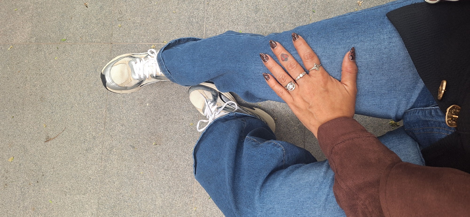 The image is a high-angle, close-up shot from a first-person perspective, featuring a person's legs, right hand, and elements of their clothing. The person is wearing loose-fitting blue denim jeans and white and silver sneakers with white laces, resting on a gray, speckled paved surface. Small pieces of dried leaves and debris are scattered across the textured, tiled ground.
The person's right hand, with an animal print (likely leopard print) nail design, rests casually on their left thigh. Several rings adorn the fingers: a band with three small stones on the middle finger, a larger circular ring with a patterned design on the index finger, and a ring with a quatrefoil-like shape on the pinky finger. A small, dark red heart tattoo is visible on the ring finger.
Part of a brown, long-sleeved garment, likely a top or sweater, is visible on the right arm. Further to the upper right, a section of a black textured garment, possibly a coat or jacket, is shown, featuring two prominent gold-colored decorative buttons.
The scene takes place outdoors during bright daylight. The ground appears to be a public walkway or square, consistent with an urban or suburban setting. The image primarily captures a casual, relaxed pose, highlighting details of personal style. No visible text or specific location cues are present within the image itself, though the provided context suggests Santa Cruz de Tenerife, Spain.