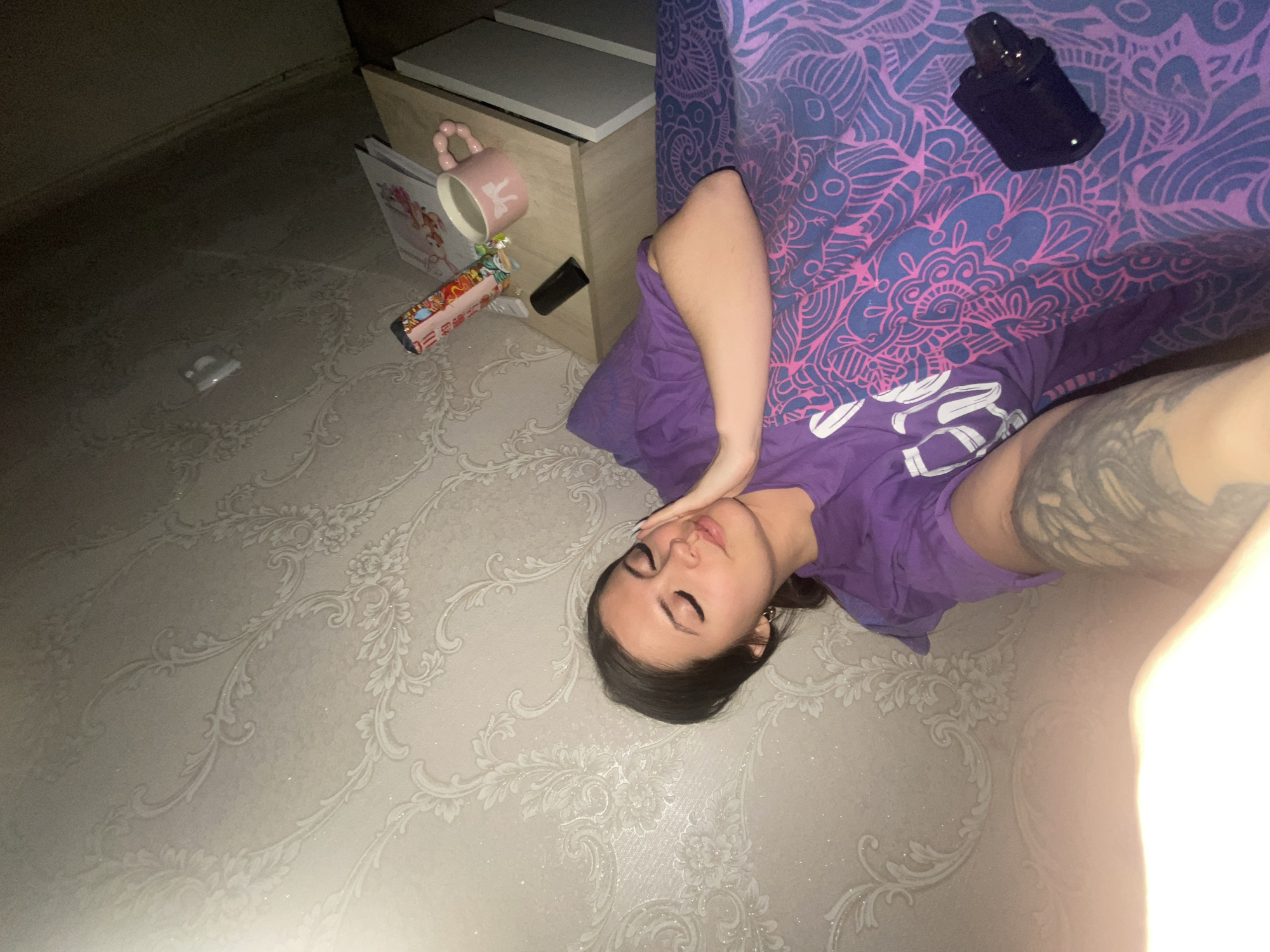 A woman with dark hair is lying on a light-colored patterned floor next to a bed. She is wearing a purple t-shirt and has a tattoo on her arm. Her hand is near her face. Next to the bed is a small nightstand with a book, a mug, and a snack on top. There is a black object on the bed. Two white pills are on the floor near the nightstand. The image was likely taken indoors in Chaikovsky, Russia.