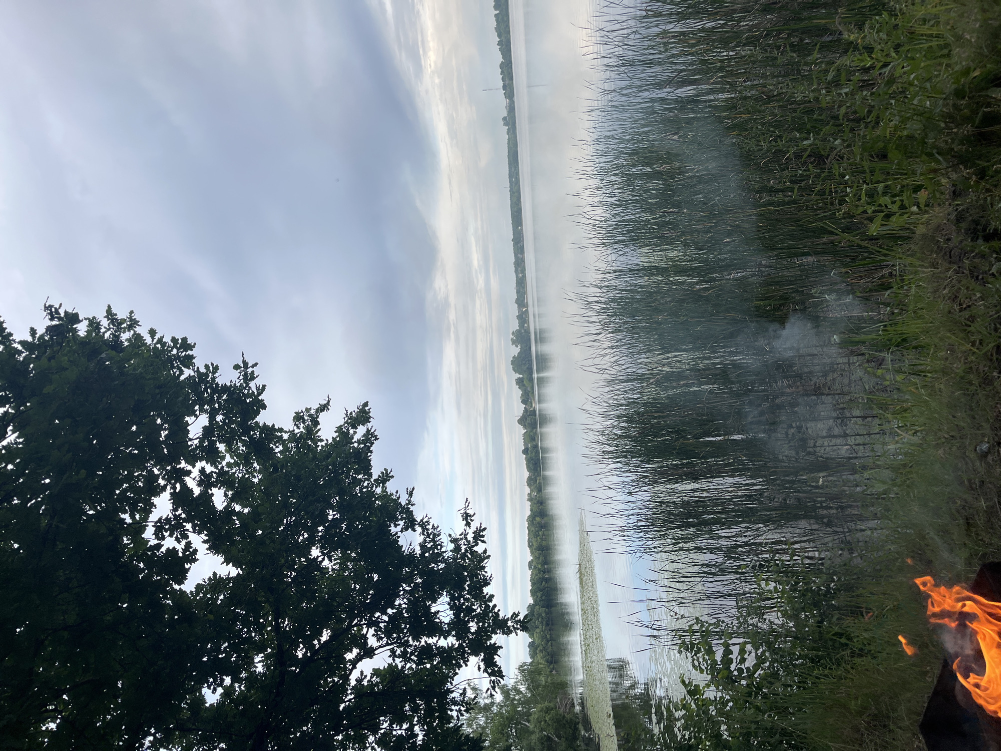 The image depicts a serene lakeside scene in Вяззеўскі сельскі Савет, Belarus. In the foreground, a bonfire crackles with vibrant orange flames, providing a warm focal point against the cooler tones of the surrounding environment. The fire is contained within a dark, unidentifiable object, likely a fire pit or grill.
To the right, tall, lush green reeds or grasses line the edge of the water, their slender stalks creating a natural barrier. A wispy trail of smoke rises from the reeds, suggesting the fire is either close to them or that something else is smoldering.
The lake itself stretches out into the distance, its surface reflecting the overcast sky. The sky is a soft blue, streaked with wispy white clouds, indicating a mild or partly cloudy day. The horizon is lined with a dense band of green trees, suggesting a forested area beyond the lake. On the far side of the lake, a thin line of what appears to be buildings or structures can be discerned, hinting at a small settlement or scattered houses.
To the left of the frame, the dark green foliage of a large tree, possibly an oak, is prominent. Its branches and leaves create a shadowy contrast with the brighter, more open sky above and the water.
The overall mood of the image is tranquil and peaceful, with the juxtaposition of the active fire and the calm natural surroundings. The time of day appears to be late afternoon or early evening, judging by the soft light and the presence of a campfire. There is no visible text in the image.