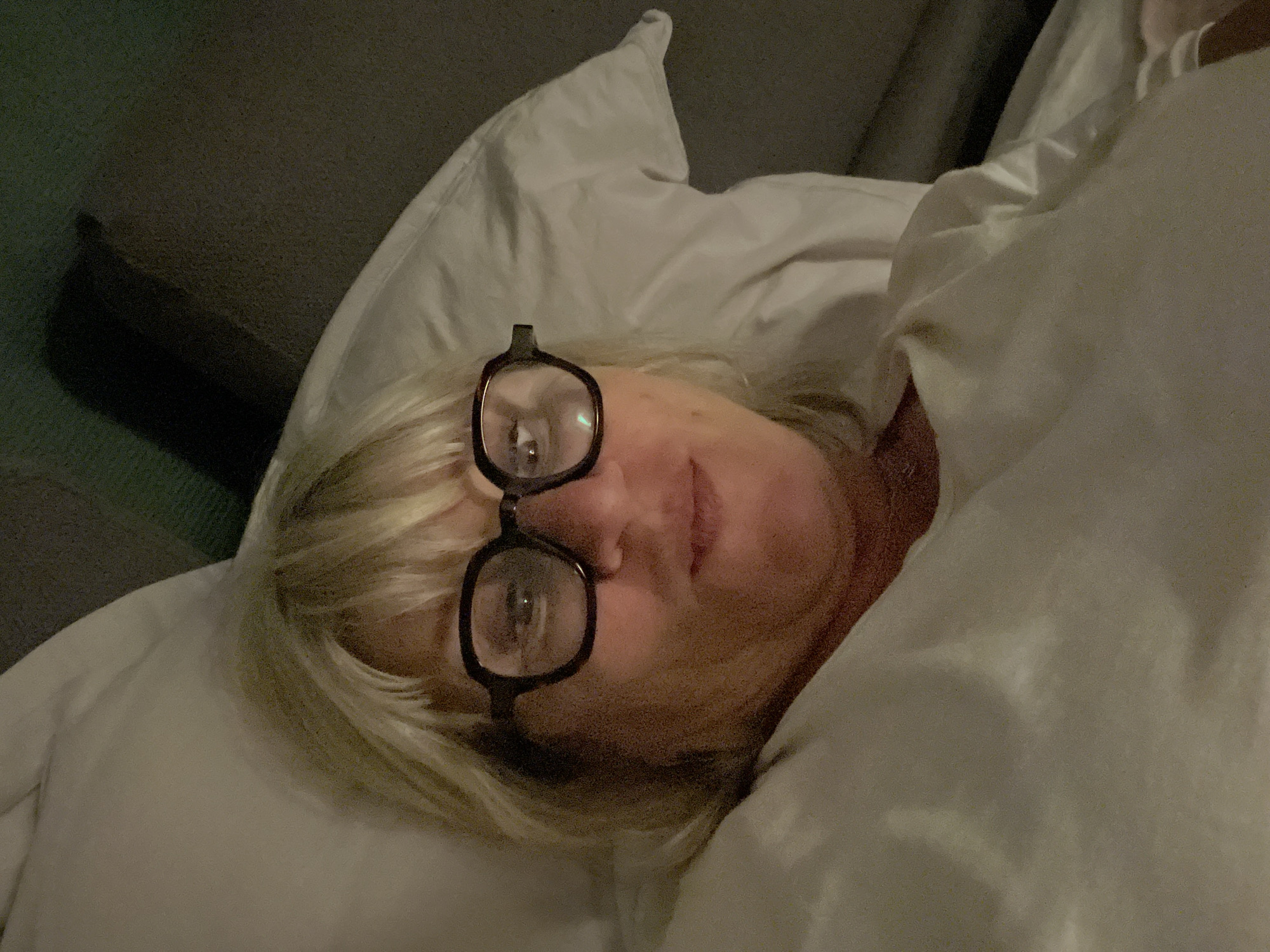 This close-up image shows a person lying in bed, partially covered by white bedding. The subject is a woman with blonde hair, which appears somewhat disheveled, suggesting a relaxed state, possibly upon waking or before sleep. She wears dark-framed, rectangular glasses that are perched slightly high on her nose and forehead, with reflections visible in the lenses. Her facial expression is neutral and calm.
She is lying on a white pillow, and a soft, white duvet or blanket covers part of her body. The overall lighting is dim, indicating it could be nighttime, early morning, or a dimly lit indoor space. The background is dark and out of focus, providing no specific details about the room beyond it being an indoor environment, likely a bedroom. The image does not contain any discernible text or specific visual cues that would directly indicate the mentioned location of Palavas-les-Flots, France. The scene conveys a sense of quiet intimacy and repose.