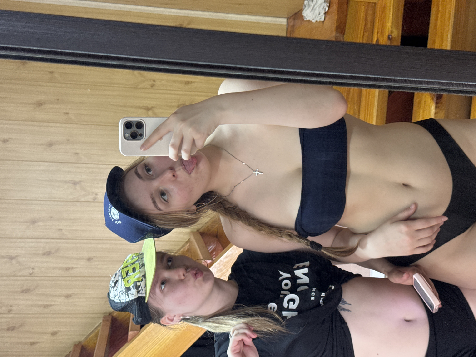 Two young women are visible in a mirror selfie. The upper woman, wearing a navy blue baseball cap and a dark blue bikini top, holds a smartphone and sticks her tongue out. She has blonde hair in a braid and is wearing a silver cross necklace. The lower woman, wearing a baseball cap with a light green brim and a black t-shirt with reversed white text, pouts her lips. She has fair skin, blonde hair, and a visible tattoo on her abdomen. Both women are wearing dark-colored bottoms. The background consists of wooden walls, a black mirror frame, and a glimpse of wooden stairs. The location is in the city of Unknown, Unknown.