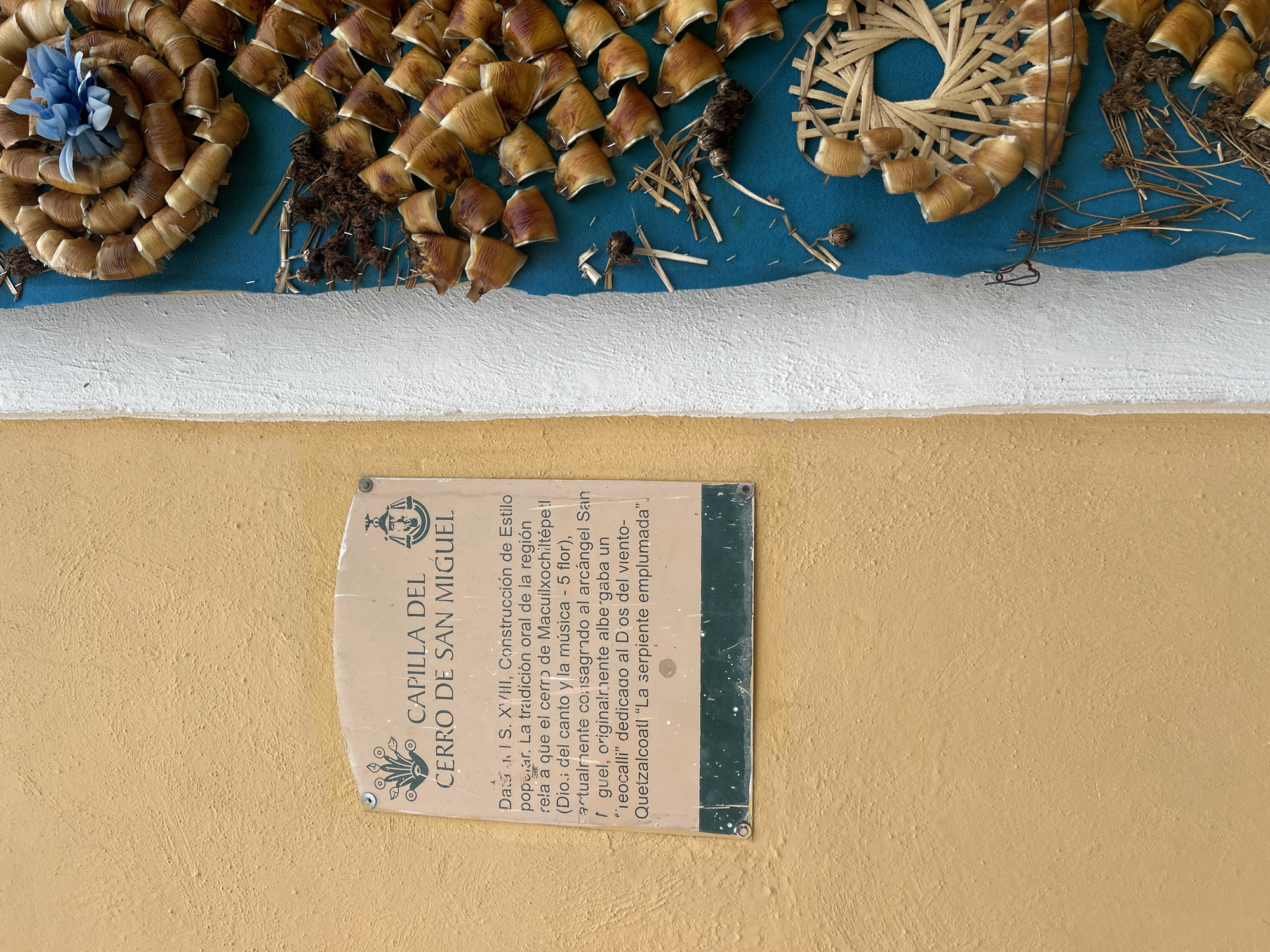 This is a close-up, eye-level, outdoor, natural light shot of a sign on a textured, mustard-yellow wall. To the right of the sign is a bright blue fabric with decorative elements made of dried plant material, possibly corn husks, arranged in circular and spiral patterns. A small blue fabric flower is attached to one of the decorative elements. The sign itself is light-colored with dark green text and a dark green border. It has a curved top edge and is attached to the wall with two visible screws. The text on the sign, in Spanish, reads: "CAPILLA DEL CERRO DE SAN MIGUEL. Data: S. XVIII, Construcción de Estilo popular. La tradición oral de la región rela a que el cerro de Macuilxochitépetl (Dios del canto y la música - 5 flor), actualmente consagrado al arcángel San Miguel, originalmente albergaba un "teocalli" dedicado al Dios del viento- Quetzalcoatl 'La serpiente emplumada'." This translates to: "CHAPEL OF SAN MIGUEL HILL. Date: 18th Century, Popular Style Construction. Oral tradition of the region relates that the hill of Macuilxochitépetl (God of song and music - 5 flower), currently dedicated to Archangel San Miguel, originally housed a 'teocalli' dedicated to the God of wind - Quetzalcoatl 'The feathered serpent'." This information indicates the location is in Atlixco, Mexico, specifically at a historical site known as San Miguel Hill.