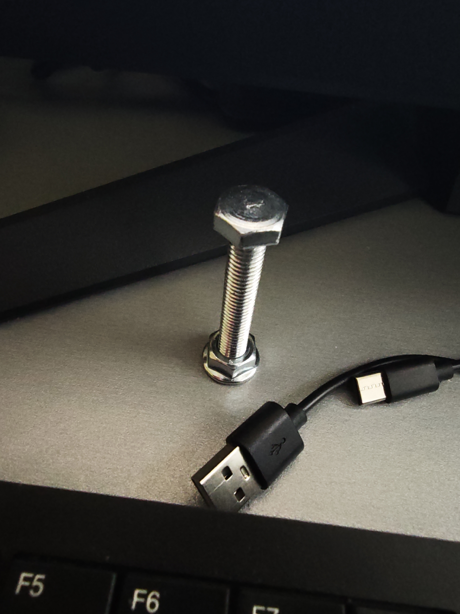 The image captures a close-up view of various items on a desk or workstation. The main subjects are a silver bolt and nut, assembled and standing upright in the center, and a black USB cable lying to its right. The bolt is hexagonal at both ends, with visible threading along its shaft, and a slight reflection of light on its metallic surface. A faint marking, possibly a '1' or a logo, is visible on the top flat surface of the bolt's head. The USB cable features a standard Type-A connector on the left, displaying the USB logo, and a Micro-B connector on the right, which has a distinct patterned metal tip.
The setting appears to be indoors, possibly an office or home office, indicated by the presence of a keyboard in the foreground and a dark, possibly electronic, object (like a monitor base or stand) in the background. The surface the items rest on is a light gray or beige material with a fine texture. Part of a black keyboard is visible at the bottom of the frame, showing the keys labeled "F5" and "F6". The lighting is even, suggesting ambient indoor light or a soft overhead source, creating subtle shadows and reflections. No specific activity is depicted, but the arrangement of objects suggests a casual placement on a workspace. The time of day is not explicitly discernable, but the lighting suggests daytime or well-lit indoor conditions. The image does not provide any visual cues to confirm the geographical location of Vasil Drumev, Bulgaria.