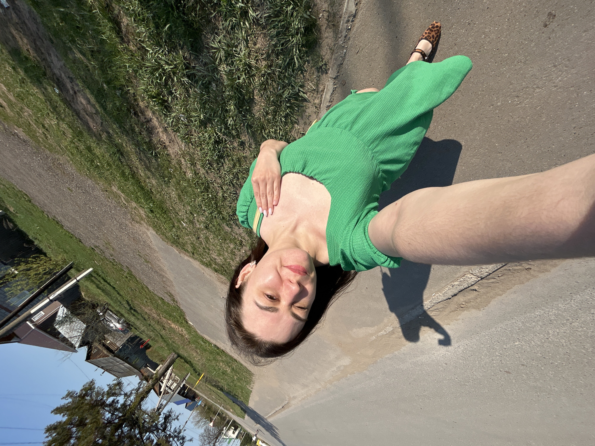 The image is a selfie taken by a woman in Chaikovsky, Russia, on a sunny day. She is standing on a paved road alongside a grassy area. She is wearing a green dress and leopard-print shoes. In the background, there are houses and trees. The image is tilted, suggesting it was taken with a hand-held device.