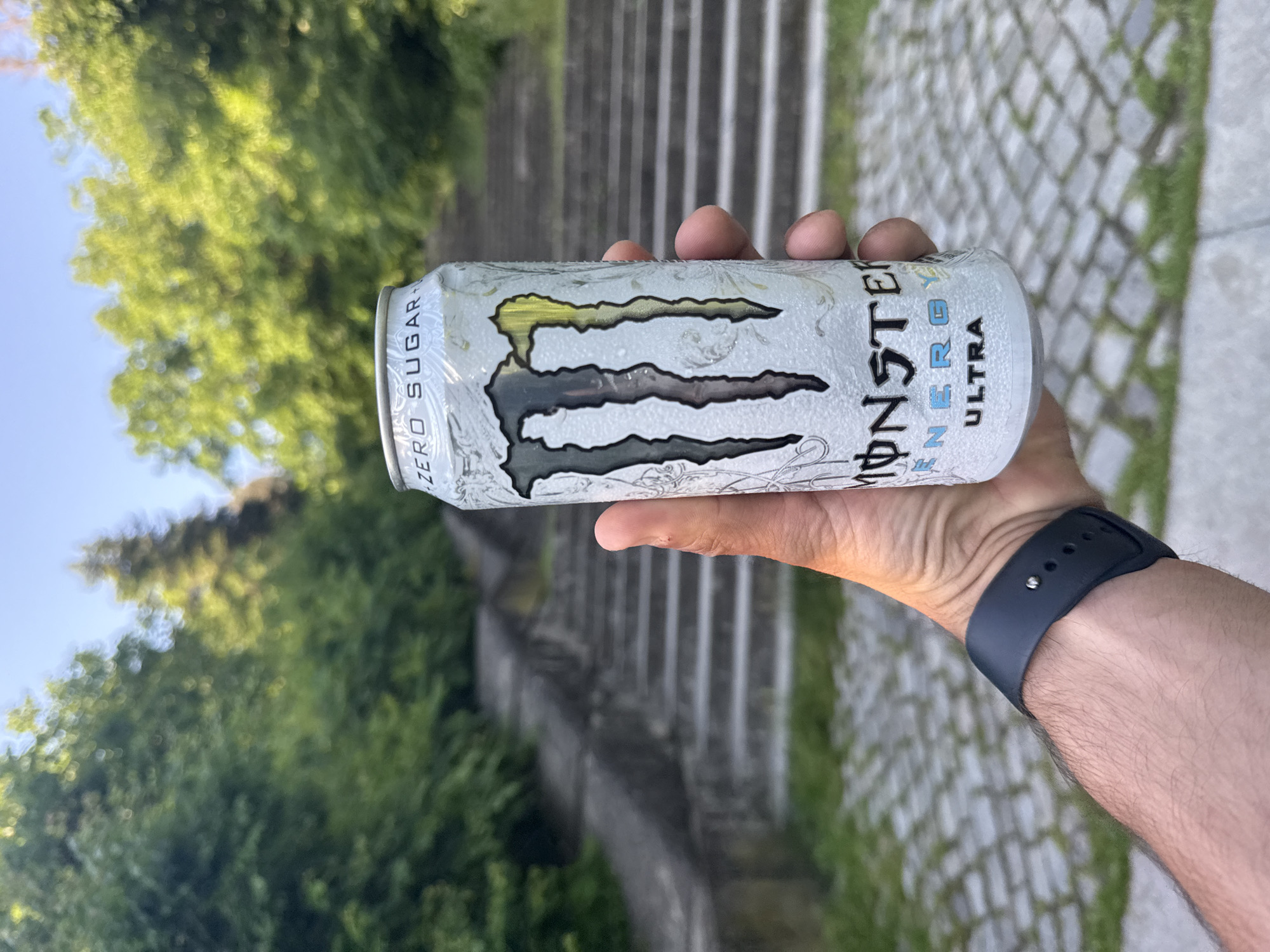 A person holds a can of Monster Energy Ultra, with the drink’s logo prominently displayed, in their right hand. The individual is wearing a black smartwatch on the same wrist. The background is a blurred outdoor scene featuring greenery, possibly trees and shrubs, and a stone structure that appears to be a staircase leading upwards. Given the location is in Shumen, Bulgaria, the staircase might be part of the Monument to 1300 Years of Bulgaria, although it's not possible to confirm this definitively based on the image alone. The day is bright and sunny.