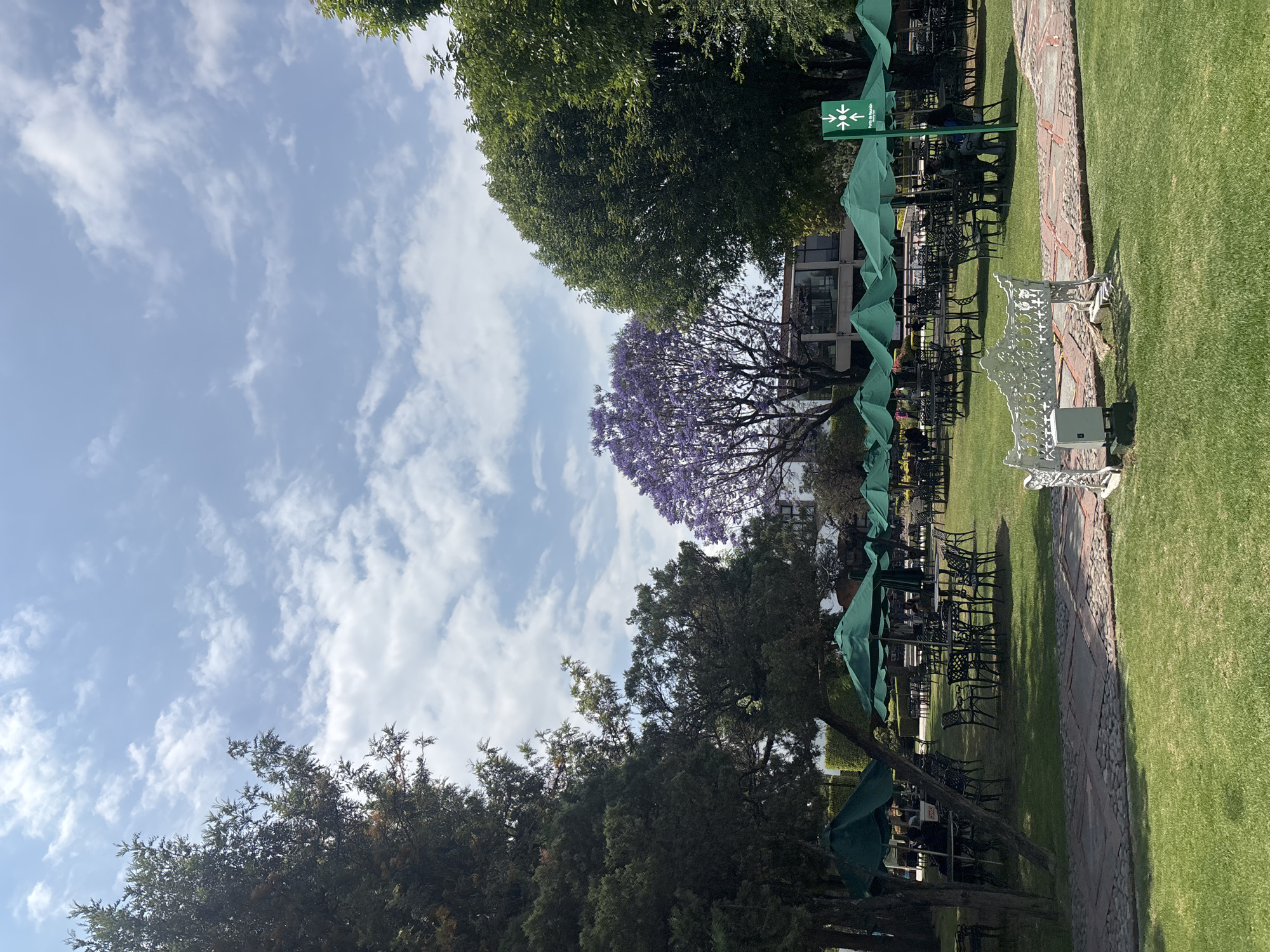 The image depicts an outdoor scene in San Andrés Cholula, Mexico, featuring a manicured lawn with a brick pathway. Several wrought-iron tables and chairs with green umbrellas are arranged for outdoor seating. A white ornamental bench is situated along the pathway. Trees, including one with purple blossoms, border the lawn. A building is visible in the background. The sky is blue with scattered clouds. No people are clearly visible.