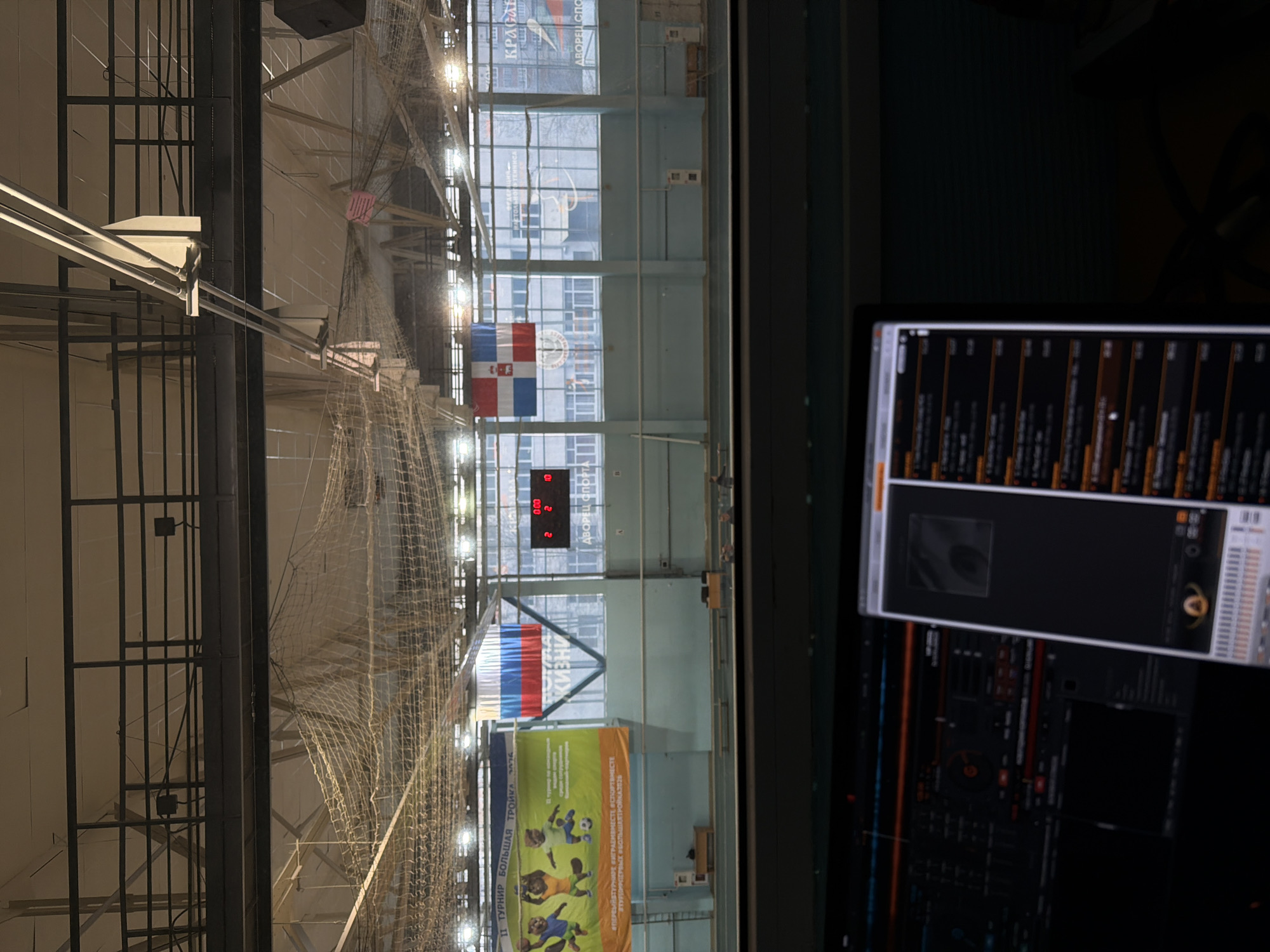 The image shows an indoor sports arena, possibly in Кондратово, Russia, based on the Cyrillic text on a banner. The arena features a high ceiling with a complex network of support beams and netting. A scoreboard displays a score of 2-2. A flag with the Russian tricolor and the name "Кондратово" on it is visible. A large banner with illustrations of children playing sports hangs on the wall. A computer monitor displaying some kind of software interface is also in view.