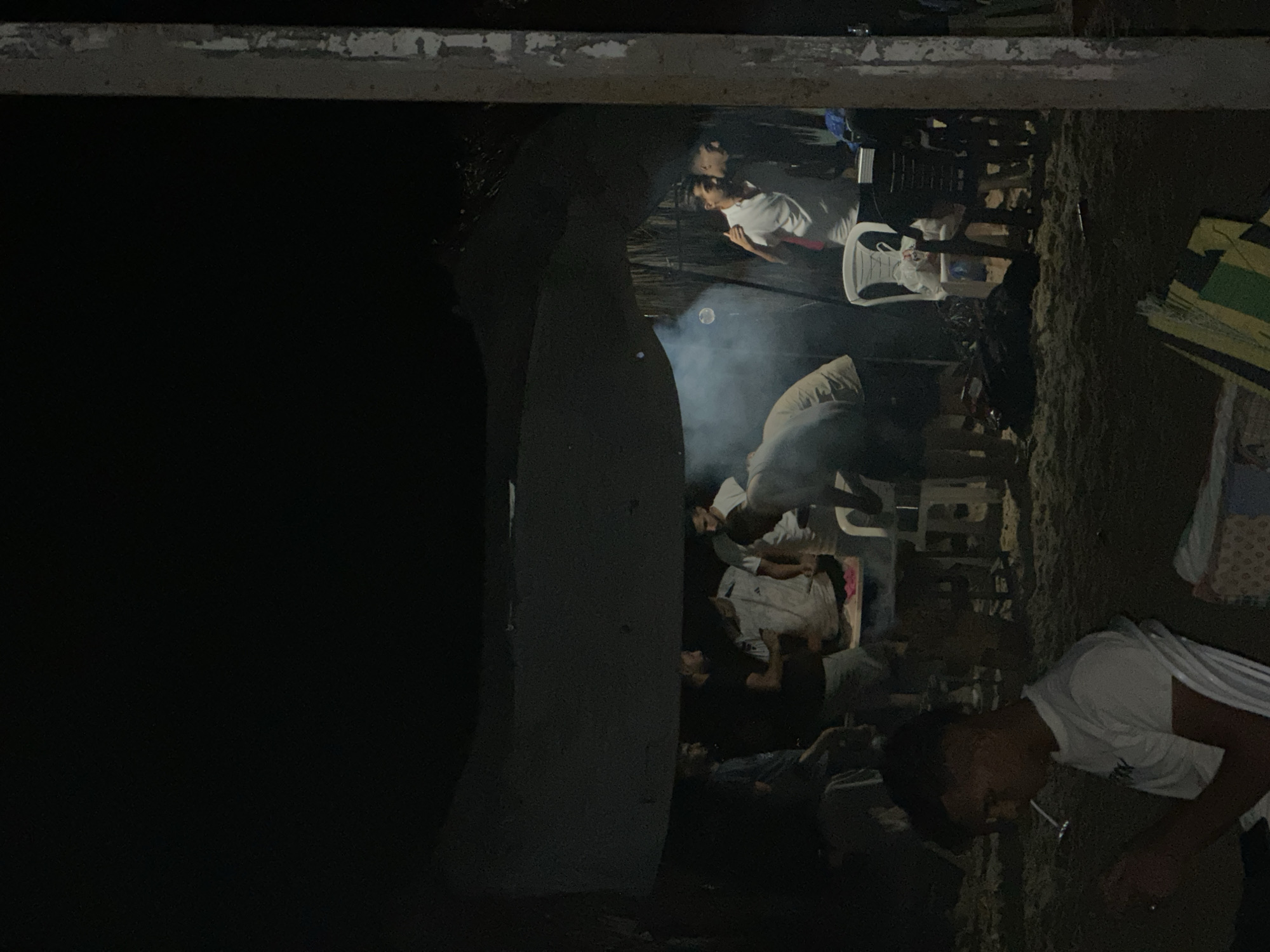 A group of men are gathered under a makeshift shelter at night, possibly on a beach. The shelter appears to be constructed from a large tarp or piece of fabric stretched between poles, with what looks like palm fronds forming a background wall. In the foreground, a young man is seated, looking down and smoking. Further into the scene, several other men are interacting, some standing around tables laden with items, and one person is seen tending to something that is producing smoke. There are chairs and other objects scattered around the sandy ground, indicating a casual gathering. The lighting is dim, with artificial light sources illuminating some of the figures and the immediate area, while the rest of the scene fades into darkness.