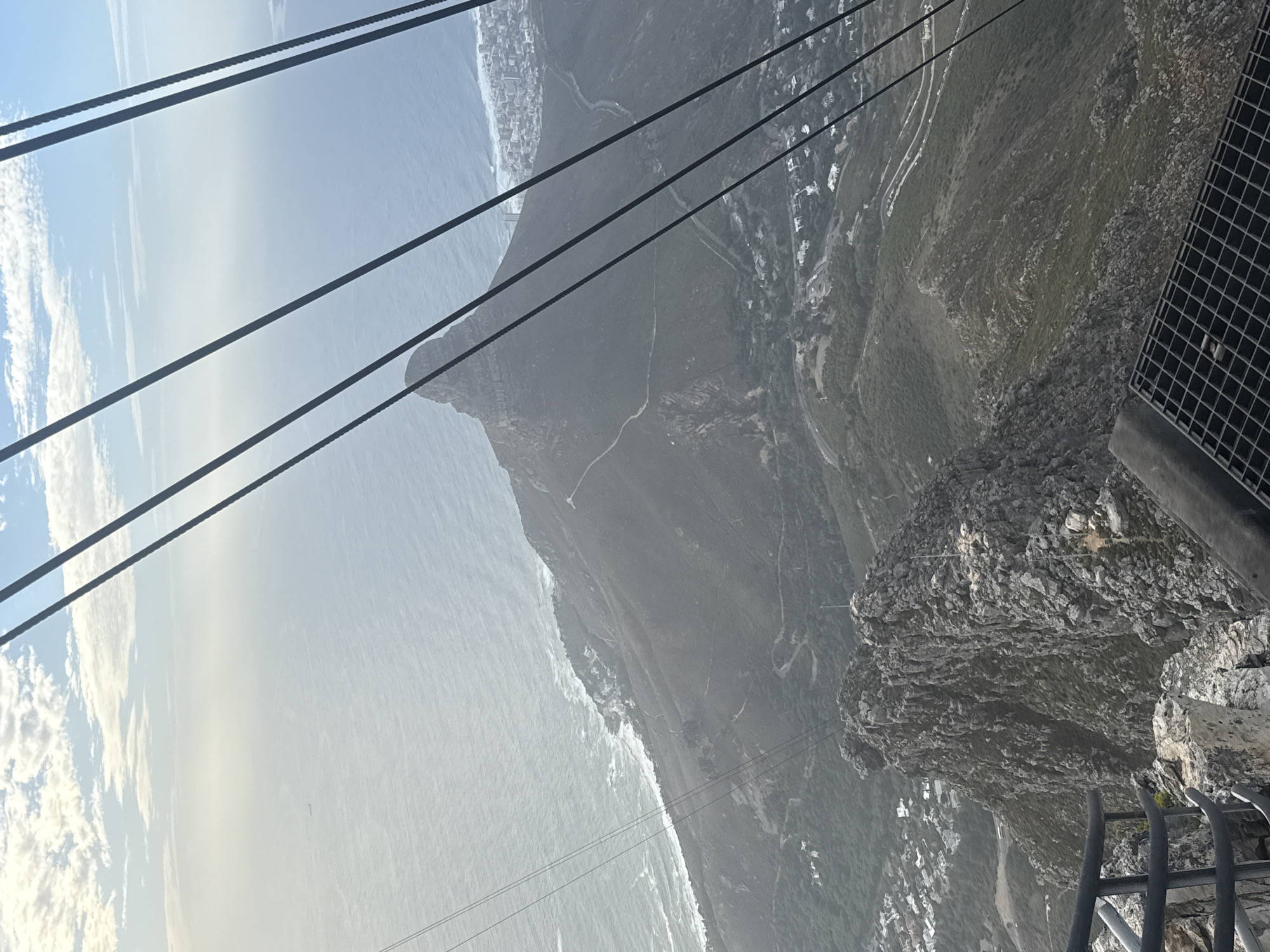 The image shows a view from a high vantage point, likely a cable car, overlooking a coastal landscape. In the foreground, several thick cables stretch across the scene from upper left to lower right. To the right is the edge of the cable car platform, with gray metal grating and a railing. The central and background of the image features the coastline of Cape Town, South Africa. To the left of the image is the ocean with white waves crashing along the coast. A prominent mountain range runs along the coast, with buildings and residential areas nestled at the base of the mountain and along the water. A road winds its way up the mountain. The overall scene is slightly hazy and the lighting is bright.