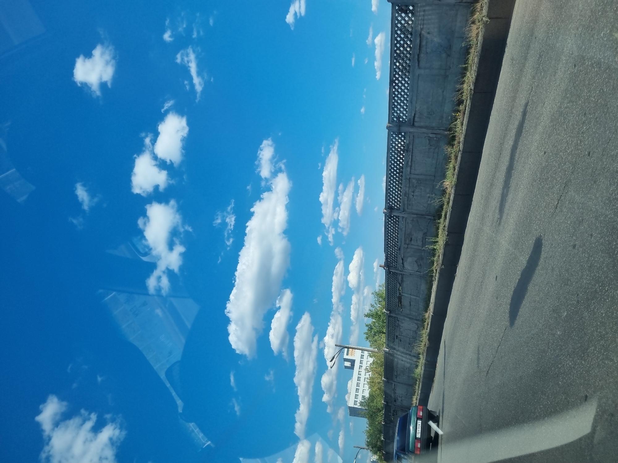 The image depicts a daytime scene from a vehicle moving along a paved road in Kyiv, Ukraine. The sky is a vibrant blue, dotted with fluffy white clouds. To the right of the road, a gray concrete barrier with a diamond-shaped pattern runs horizontally. Behind the barrier, lush green trees are visible, and beyond them, a white multi-story building with numerous windows stands under the sky. A single lamppost is situated between the trees and the building. On the road in the foreground, the rear of a dark green car is visible, facing away from the viewer, with its license plate displaying "151-93KT". The road itself shows some cracks and is bordered by a concrete curb and some sparse vegetation. The scene appears to be an urban or suburban environment.
