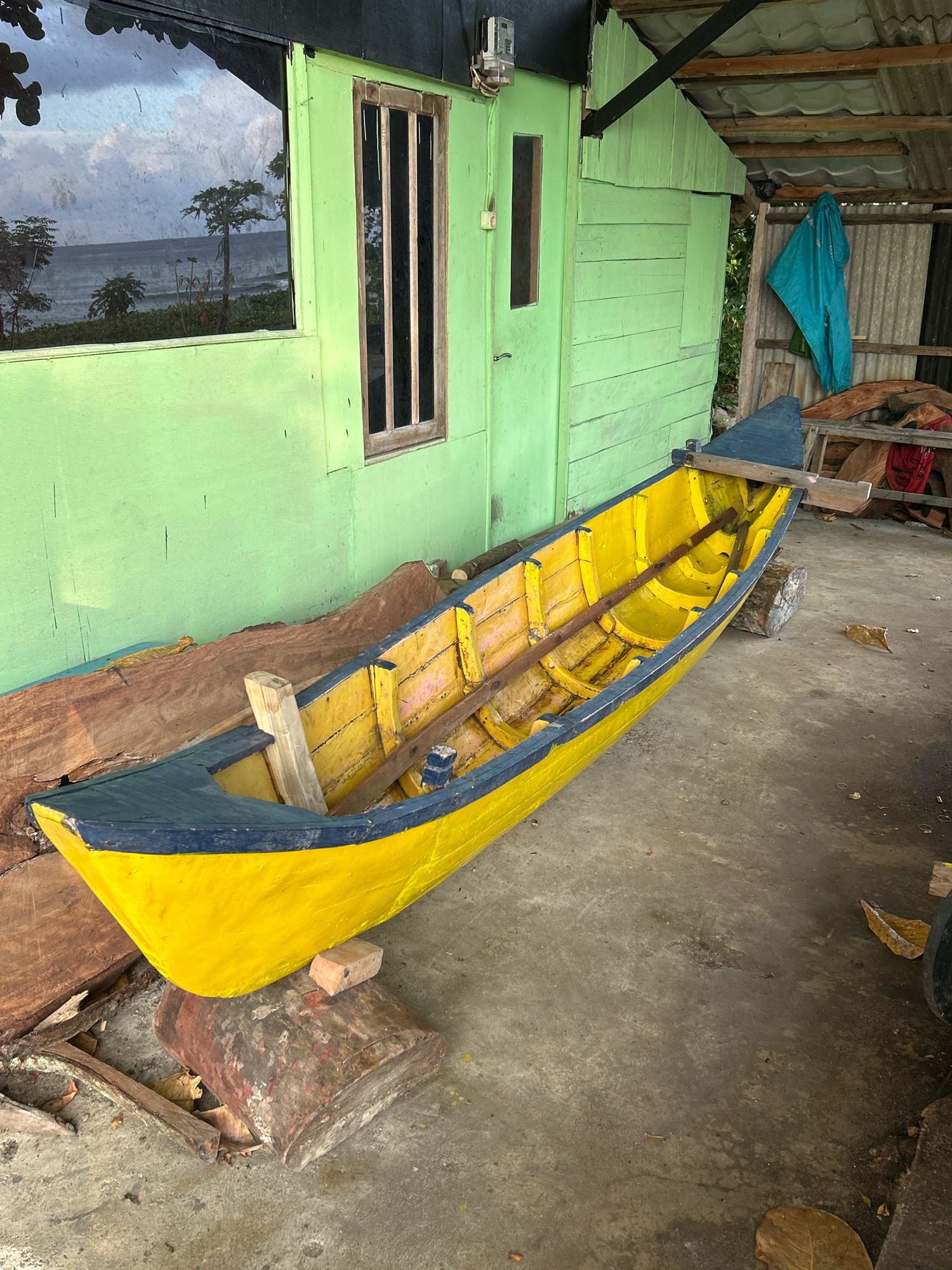 In Sabang, Indonesia, a yellow and blue wooden boat rests on a concrete floor, propped up by logs and a brick. The boat is in the foreground, angled diagonally. Behind it, a bright green wooden structure with a dark-framed window and a closed door stands. A blue tarp hangs from the roof of an open-sided shelter to the right of the green building. The scene suggests a workshop or storage area, possibly near a coastal environment, as indicated by the reflection of trees and the sea in the window of the green building.