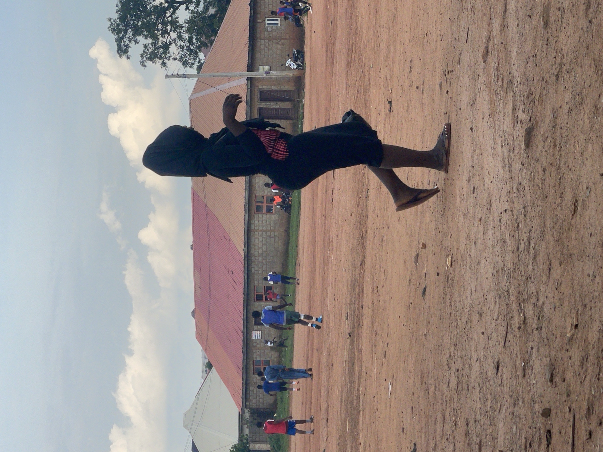 In Kurunduma, Nigeria, a woman wearing a black hijab and a black outfit with a striped undershirt walks across a dusty, outdoor sports field. The field is composed of reddish-brown dirt and scattered debris. In the background, a low-rise building with a red corrugated metal roof and light-colored brick walls is visible. Several individuals are scattered across the field, appearing to be engaged in a game of soccer. One person in a red shirt and blue shorts stands on the left side of the frame, while another in a blue vest and green shorts is near the center. A few other figures in athletic attire are also present, some running or standing. A tree with green foliage stands to the right of the building, and power lines extend across the sky. The sky is partly cloudy with white clouds against a light blue backdrop. A motorcycle is parked near the building, and some individuals are seated nearby.