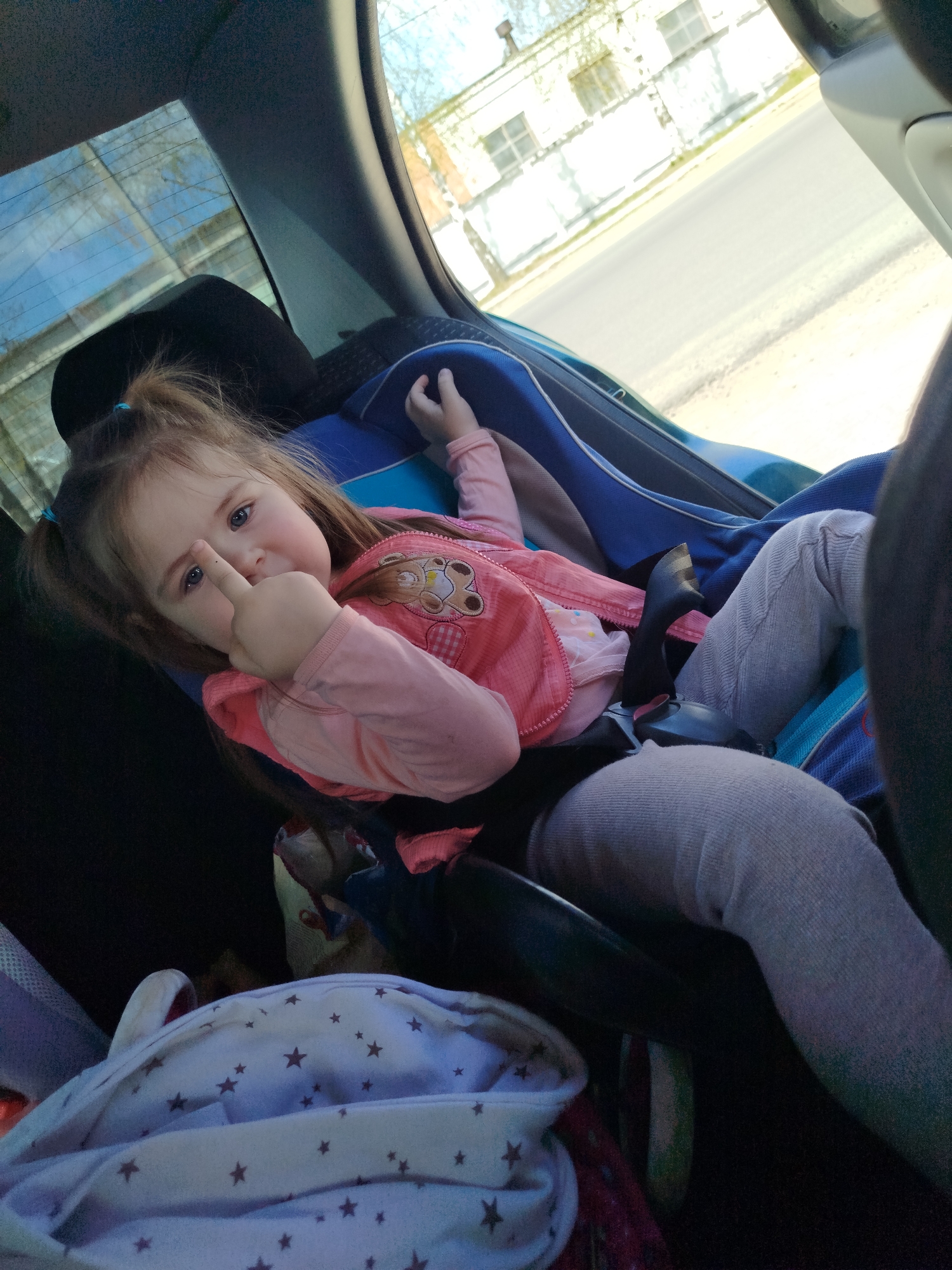 A photograph shows a young girl, approximately 3-5 years old, secured in a child safety seat in the back of a vehicle. She is wearing a pink jacket over a light pink shirt and gray leggings. Her brown hair is styled in pigtails with blue hair ties. The girl is looking directly at the camera and extending her middle finger upwards towards her forehead. She is secured by the car seat's harness.
Visible in the background through the car window is a street scene with buildings. The scene appears to be a residential or commercial area. The location can be identified as Chaikovsky, Russia. A white backpack with gray stars and a dark seat are also present.