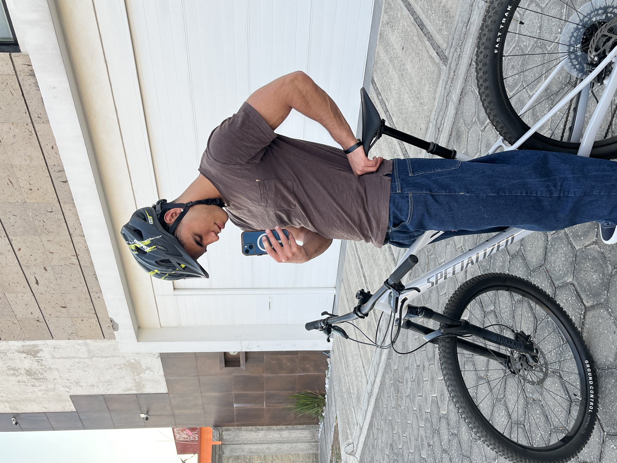 The image shows a man standing next to a mountain bike in San Andrés Cholula, Mexico. He is wearing a helmet, a brown t-shirt, and blue jeans. He is holding a blue smartphone and appears to be taking a picture. The bike is white with black tires. The background includes a light-colored building with horizontal siding and a stone tile walkway.