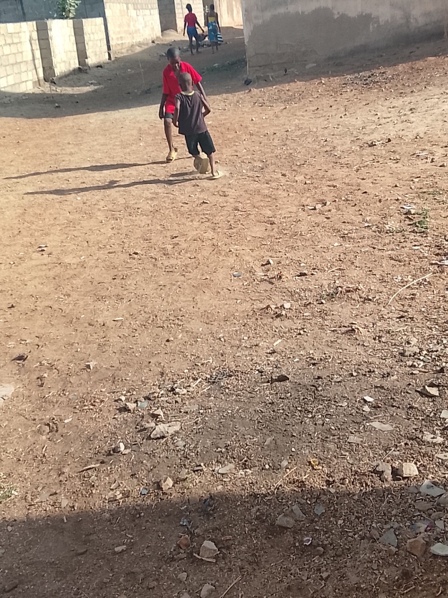 This image captures a vibrant scene of children playing football (soccer) on a dusty, unpaved ground in Bauchi, Nigeria. The main subjects are two young boys. The boy in the foreground, wearing a dark shirt, shorts, and flip-flops, is actively dribbling a makeshift ball, which appears to be a stone or a bundled object, with his right foot. His posture suggests focus on the game. Behind him and slightly to his left, another boy in a red short-sleeved shirt and shorts is also engaged in the play, possibly chasing the ball or observing.
In the background, several other individuals are visible, walking past simple, unpainted block buildings. One person is dressed in a red top and blue shorts, while another wears a striped garment. The setting is an open, sun-drenched area, likely a community space or an unpaved street. The ground is composed of hard-packed earth, scattered with small rocks and debris, characteristic of arid or semi-arid environments.
Notable details include the long, distinct shadows cast by the children and buildings, indicating that it is either early morning or late afternoon. The clear blue sky and bright sunlight suggest a dry, sunny day. The scene portrays a slice of everyday life, highlighting the simple joy of childhood play amidst modest surroundings. There is no visible text within the image.