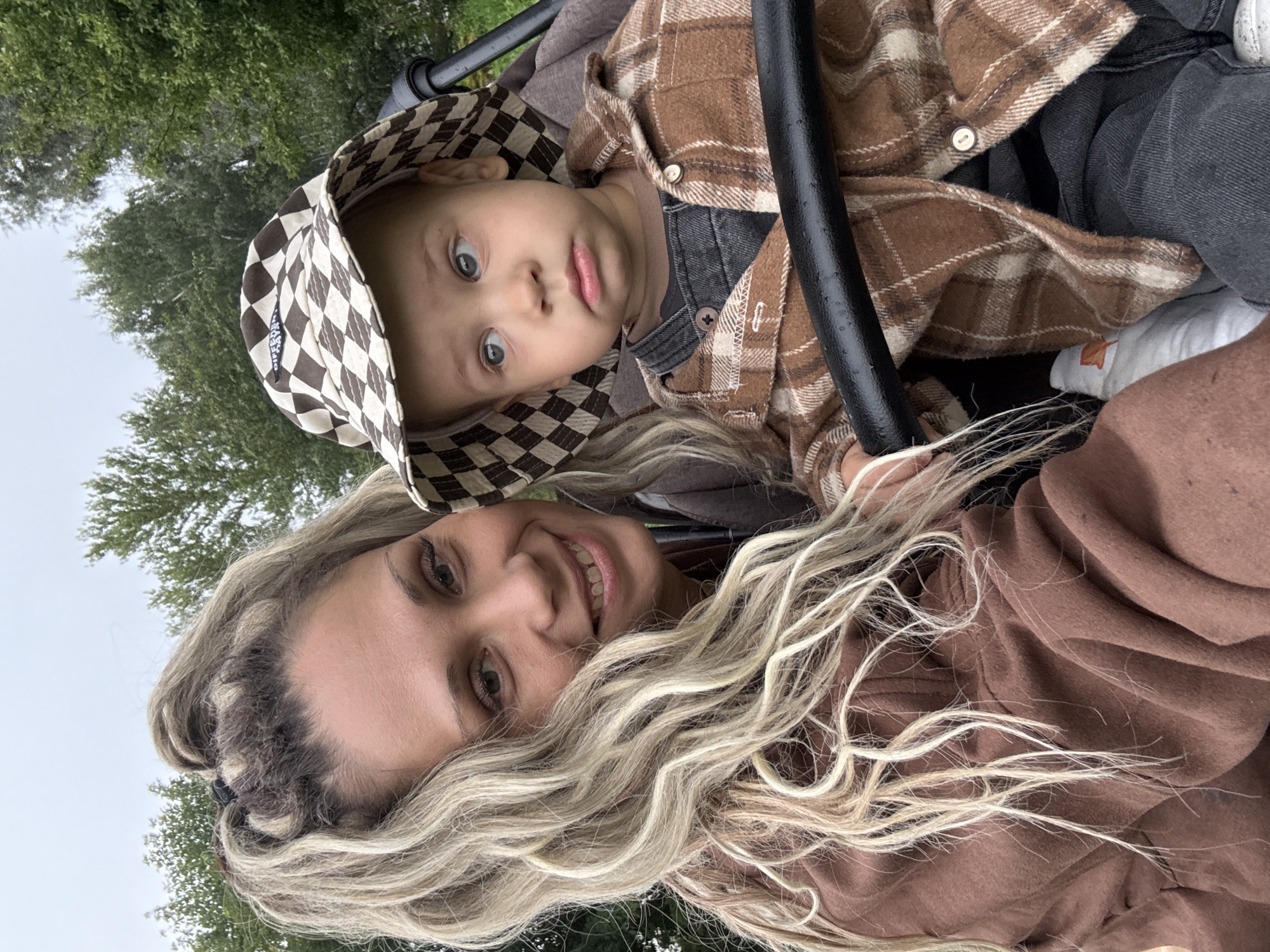 In Rusanovo, Russia, a woman with blonde hair pulled into a high ponytail and wearing a brown hoodie is taking a selfie with a young child in a stroller. The child, wearing a checkered bucket hat and a brown plaid shirt over dark overalls, looks to the side with a slightly pensive expression. The background consists of lush green trees under an overcast sky, suggesting an outdoor setting. The woman is smiling and looking at the camera, while the child is looking away, creating a contrast in their gazes. The stroller's black handlebar is visible in the foreground, framing the subjects.