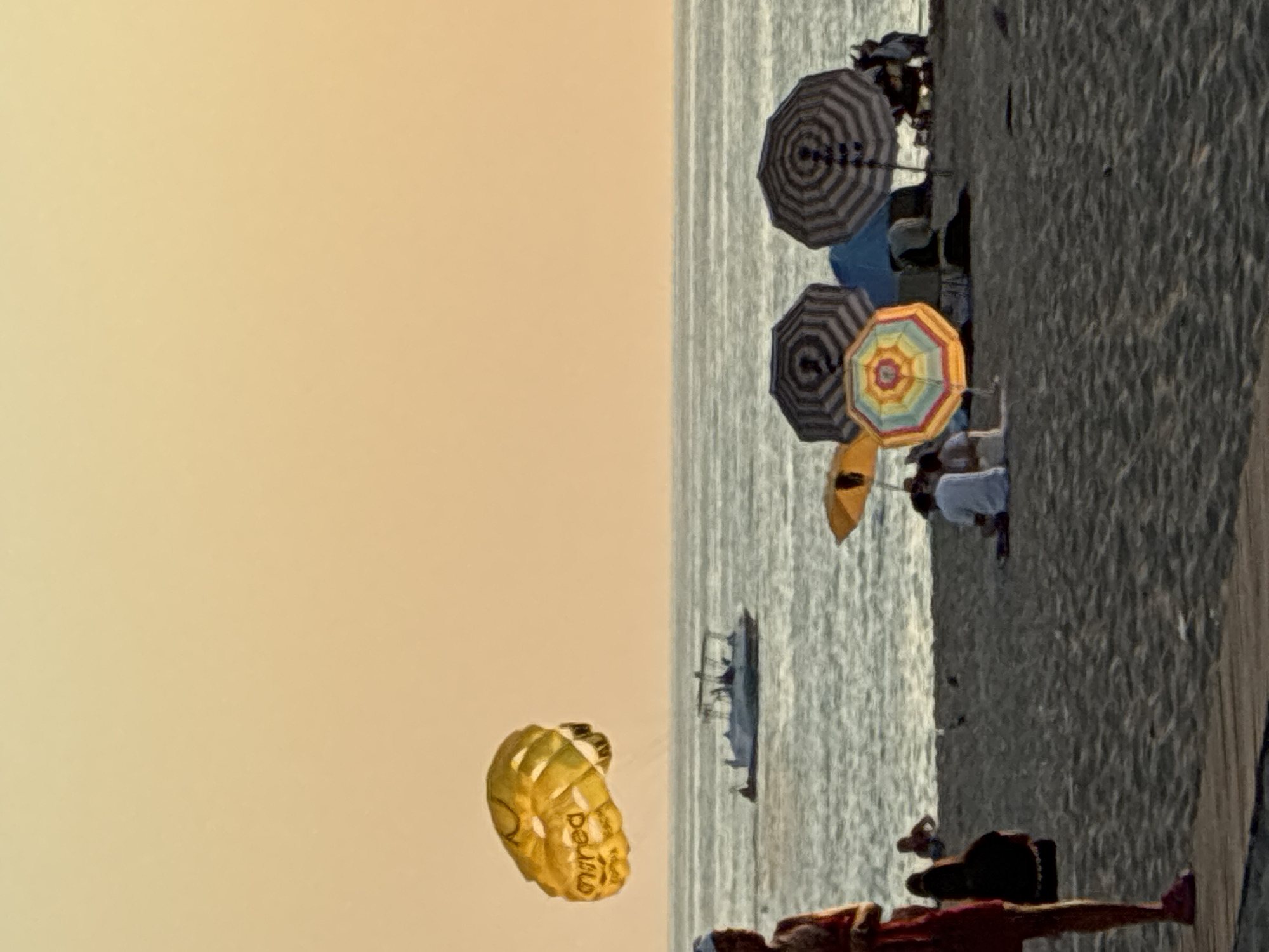 This image depicts a beach scene in Alanya, Turkey, during a sunset. A bright yellow parasail is visible in the sky, with some text that appears to be in a foreign language. Below the parasail, a boat is cruising on the water. On the sandy beach, several people are gathered under various colorful umbrellas, some sitting and others standing. One person in the foreground, on the left side of the image, is wearing a red dress and pink shoes. The sky is a soft, warm orange, indicating the time of day. The water is a muted blue-grey, and the sand is a dark, textured brown.