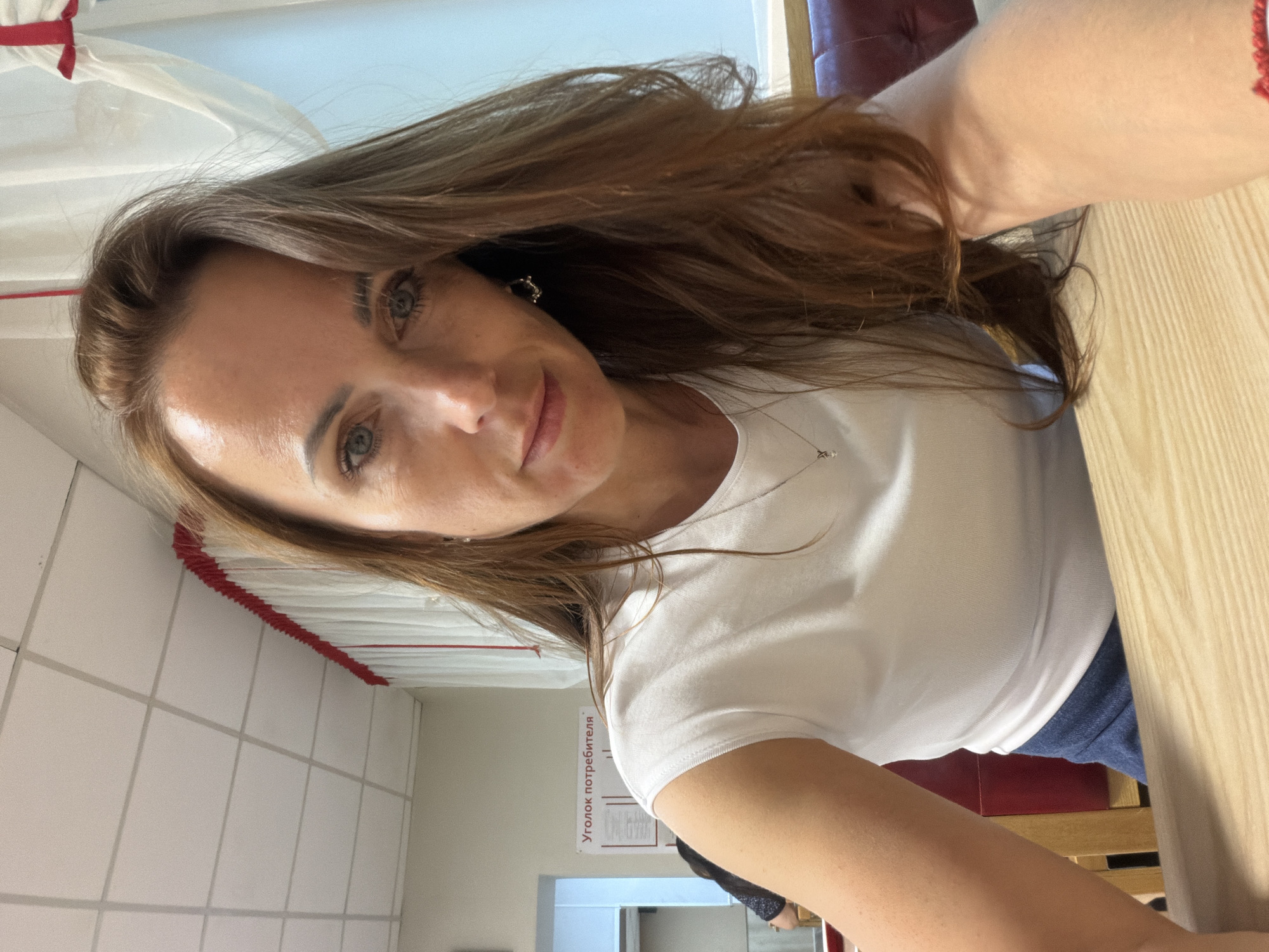 A woman with long brown hair is seated at a wooden table, looking directly at the camera. She is wearing a white t-shirt and a dark blue skirt. In the background, there is a white curtain with red trim, a sign that reads "Уголок потребителя" (Consumer's Corner), and other patrons seated at tables. The setting appears to be a cafe or restaurant.