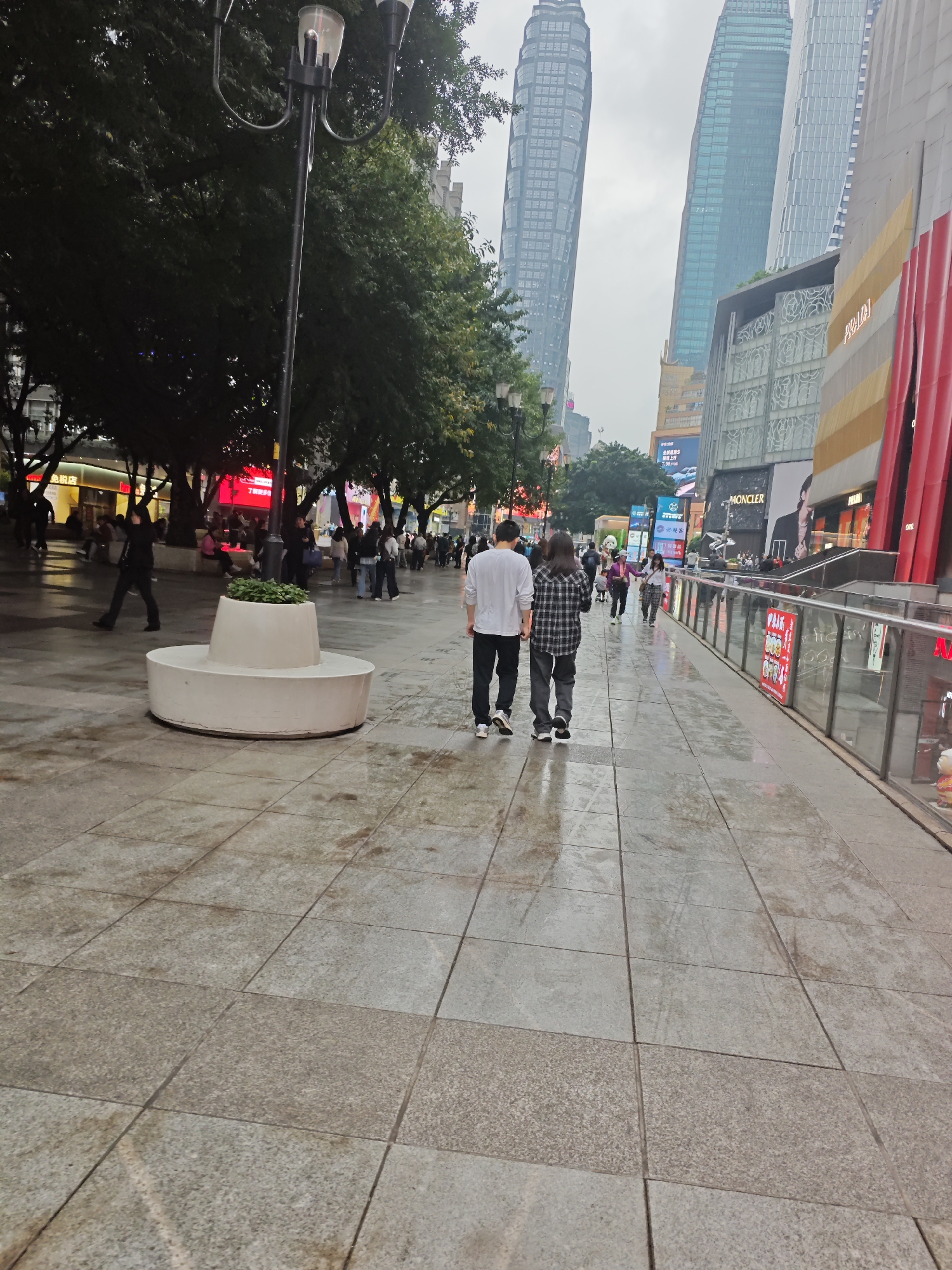 A slightly high-angle view captures a wet, paved pedestrian area in Yuzhong District, China. Two people, a male in a white shirt and black pants and a female in a plaid shirt and gray pants, are walking away from the viewer. Buildings, including skyscrapers and retail spaces with large advertisements (PRADA, MONCLER), flank the pedestrian area. Other pedestrians are visible in the background. A white circular planter sits on the left. Lamp posts line the street. The sky is overcast.