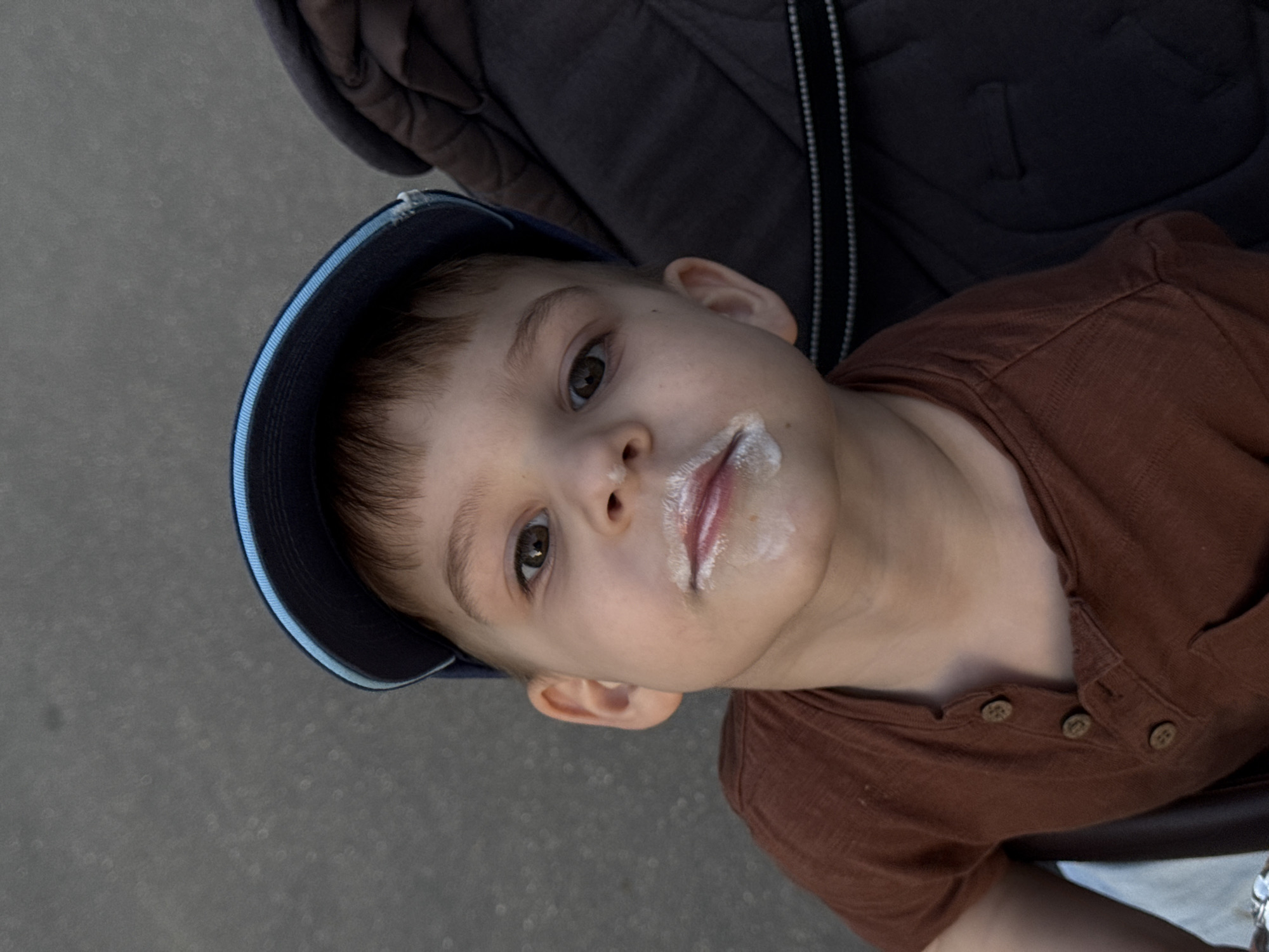 A young boy with white cream residue around his mouth, possibly from ice cream or yogurt, gazes upward. He wears a blue baseball cap and a brown, button-down shirt. The boy appears to be seated in a stroller or some sort of child carrier, suggested by the dark, textured fabric behind him. The location is not determinable from this image.
