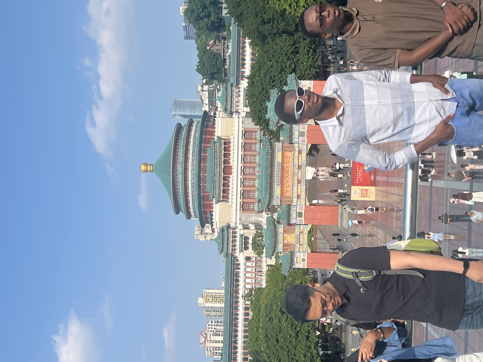In Yuzhong District, China, under a bright blue sky with scattered white clouds, stands the Great Hall of the People. This iconic building features a prominent green, multi-tiered roof topped with a golden spire, and a wide, white facade with red accents. In front of the building, a large plaza is visible, filled with numerous people walking and gathering. Verdant trees surround the area, with modern high-rise buildings in the background. In the foreground, three young men are positioned on a raised platform with a railing. The man on the left, wearing glasses, a black polo shirt, and jeans, carries a backpack. The man in the middle, sporting white sunglasses and a striped long-sleeved shirt, has his hands in his pockets. To the right, a man wearing glasses and a brown striped short-sleeved shirt stands with his hands clasped. The overall scene depicts a bustling urban environment with historical architecture.