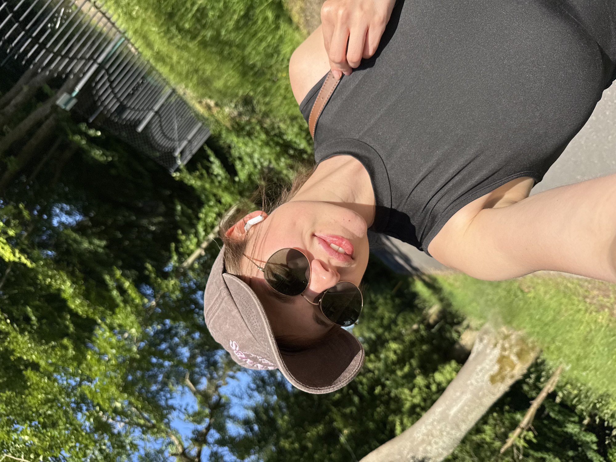 A young woman is taking a selfie outdoors, wearing a brown baseball cap, sunglasses, and a black sleeveless top. She has white wireless earbuds in her ears and a brown strap from a bag is visible over her shoulder. She is smiling, with her head tilted slightly. The background shows a park-like setting with green trees, grass, and a fence. There is a fallen tree trunk visible on the left side of the image. The sunlight suggests it is a bright, clear day. The location is identified as Szklarska Poręba, Poland.