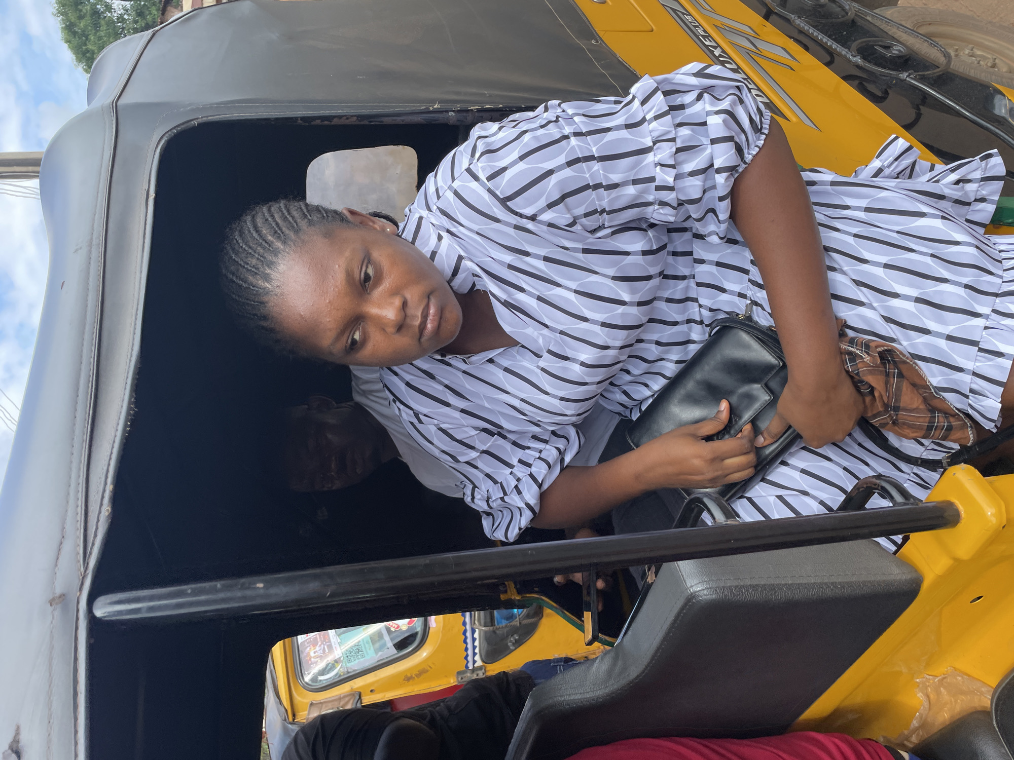 In Nkpor, Nigeria, a young woman with braided hair, wearing a black and white striped dress with ruffled sleeves, sits in the passenger seat of a yellow auto-rickshaw. She holds a black clutch bag and a brown plaid cloth in her lap. Another passenger, a man in a white shirt, is visible in the driver's seat. The auto-rickshaw has a black roof and a black interior. The scene takes place outdoors, with a cloudy sky and glimpses of trees and buildings in the background.
