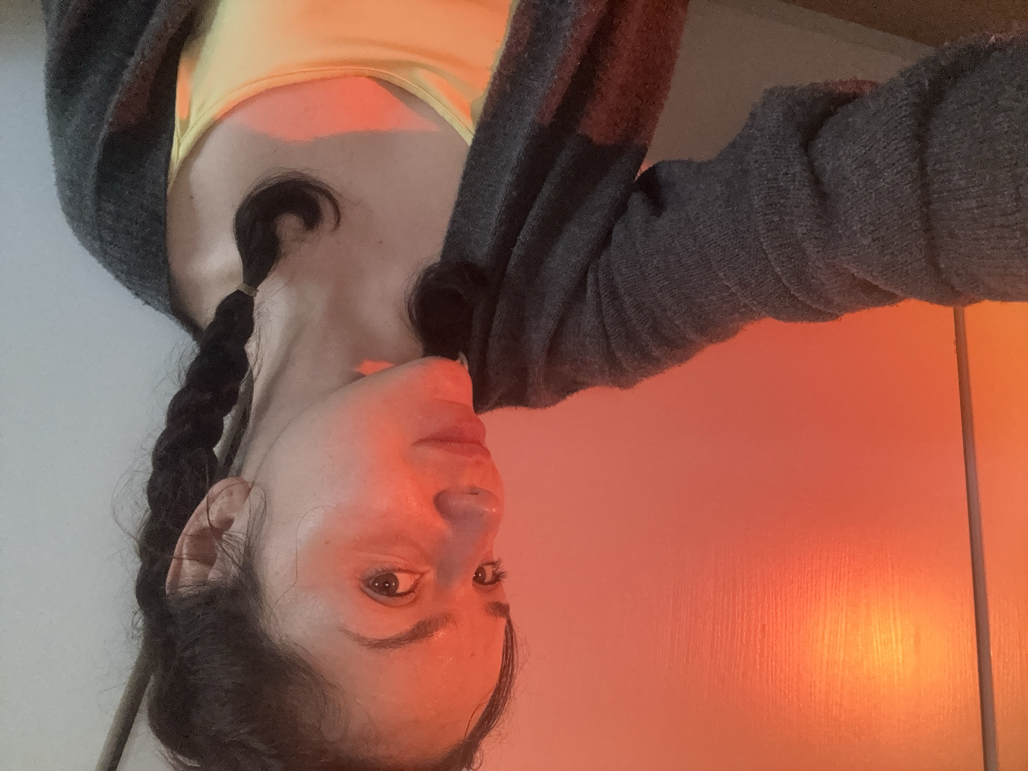A woman with dark hair braided on both sides is shown in a portrait-style image, oriented upside down. She wears a yellow top and a grey cardigan. The background appears to be a light-colored wall on the left and an orange-toned surface with vertical lines on the right. The face is illuminated with a reddish tint. The location is not determinable from this image.