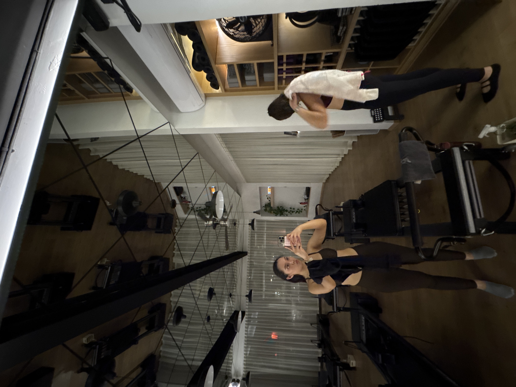 The image depicts a person in a yoga studio in Puebla, Mexico. The studio features pilates equipment and a mirrored ceiling, which reflects the person taking a selfie. Other details in the scene include shelving units and decor, suggesting a commercial or specialized space for physical activities.