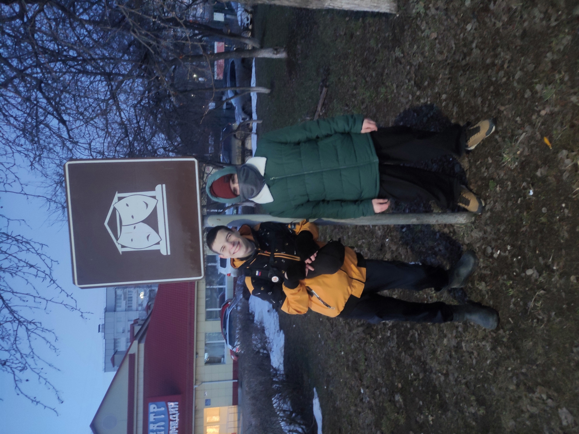 Two people, one wearing a green puffer jacket with a hood and the other wearing an orange and black jacket, are standing near a brown sign with a white theatrical mask symbol. The sign, likely indicating the location of a theater, is situated in front of a building with red and beige facade, and a sign in Russian that includes the word "ЦТР". A bare tree is visible in the background against a twilight sky. The location is likely in the city of Chaikovsky, Russia, based on the Cyrillic lettering on the building.
