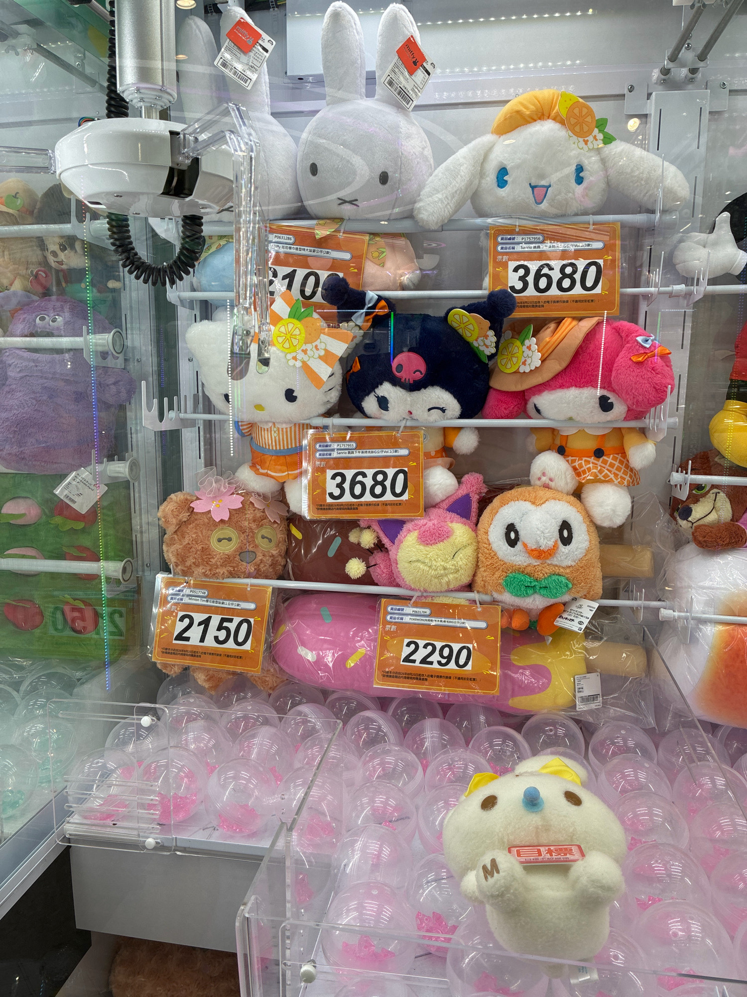 The image shows a claw machine filled with plush toys. In the foreground, a white plush toy with a blue nose and yellow ears is visible. Behind it, a collection of clear plastic spheres contains pink objects. In the middle ground, various plush toys are displayed on shelves, including a Hello Kitty plush, a pink bunny plush, a brown bear plush with a pink flower, an orange owl plush with a green bow, and a black plush with a pink skull. There are also larger plush toys visible in the background, such as a white bunny and a white character with an orange hat and fruit accents. Price tags with numbers like "2150", "2290", and "3680" are attached to some of the toys. The claw mechanism of the machine is visible at the top. The overall environment suggests a retail or entertainment setting, likely a shopping mall or arcade, with the specific location being Hong Kong, China, based on the signage and typical arcade establishments found there.