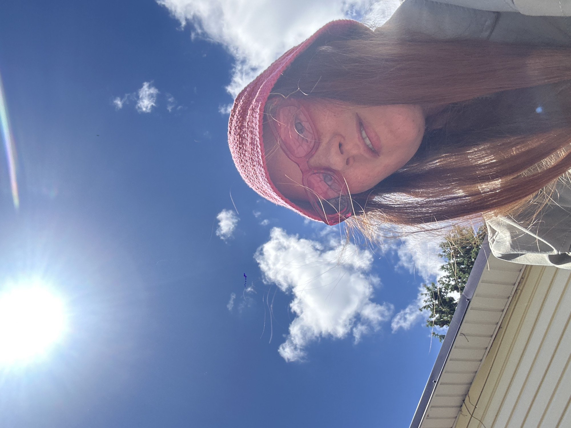 The image, captured in Nizhny Novgorod, Russia, presents a low-angle view looking up towards a person and the sky. The subject, likely female, is looking directly down into the camera with a slight smile or smirk, indicating a casual and relaxed demeanor. She wears a pink knitted hood or beanie and prominent pink, round-rimmed glasses. Her long, reddish-brown hair frames her face and flows downwards, with several strands visible against the bright sky.
The background features a vivid blue sky dotted with scattered white, fluffy clouds. A brilliant sun occupies the lower-left corner, causing significant lens flare and bright illumination. On the right side of the frame, a portion of a building with light-colored siding and a grey gutter or eave is visible, along with the foliage of a green coniferous tree. Several fine, wispy strands or lines can be seen in the sky, possibly stray hairs or environmental elements. A tiny dark speck, potentially a bird or insect, is also faintly discernible against the blue expanse. The overall scene suggests a sunny outdoor setting during the day. No visible text is present.