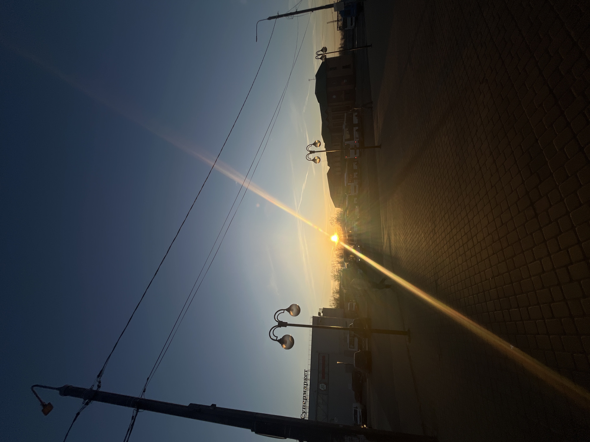 The image depicts an urban outdoor scene in the city of Klin, Russia, during either sunrise or sunset, characterized by a low sun angle and warm illumination. The sky is clear, transitioning from deep blue overhead to orange and yellow at the horizon, with a prominent diagonal lens flare spanning from the lower-left to the upper-right of the frame.
In the immediate foreground, a paved surface composed of dark, rectangular interlocking pavers is visible, partially illuminated by the direct sunlight and showing lens flare.
The midground features several elements:
* Multiple human silhouettes are discernible, walking or standing, primarily obscured by backlighting and lens flare.
* Two decorative streetlights, each with two spherical lamps and curved support arms, are positioned centrally. Another simpler streetlight is visible on the far right.
* A utility pole with multiple overhead wires runs vertically on the far left, with additional wires stretching horizontally across the upper frame.
In the background, two prominent buildings define the cityscape:
* To the left, a large, light-colored building with multiple windows features the word "Супермаркет" (Supermarket) on its upper facade. Several vehicles, including white and dark cars, are parked or moving in front of it.
* To the right, a darker-colored, multi-story building with a distinctive green-peaked roof stands prominently. Multiple large white buses are parked in front of this building.
* A line of bare trees forms a dark silhouette against the horizon between the buildings. The sun itself is a bright, intense point of light near the center-right horizon.