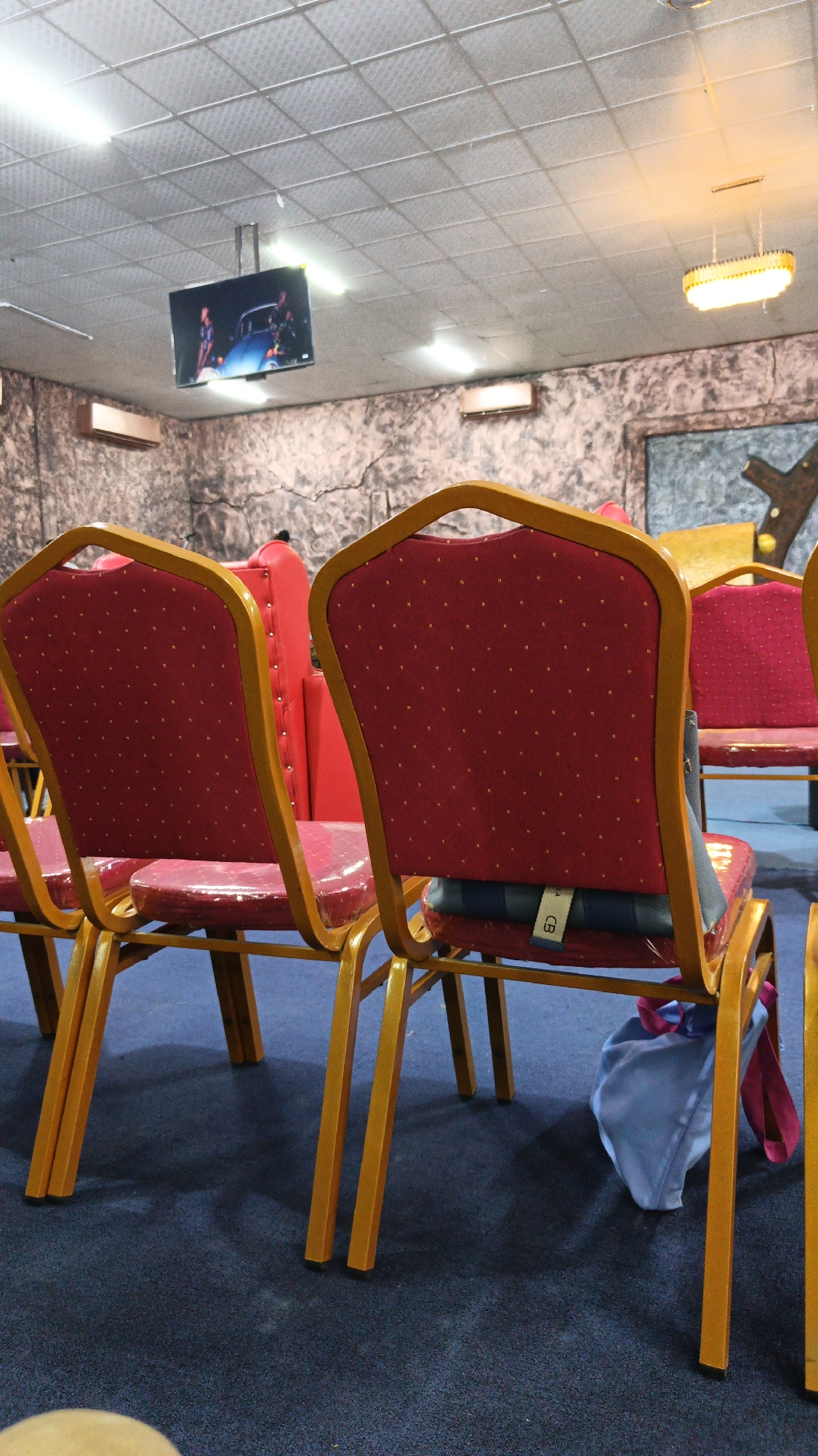The image captures the interior of a room, possibly a conference hall or a place of worship, in Dar es Salaam, Tanzania. The foreground is dominated by rows of red upholstered chairs with gold-colored frames. The chairs are arranged in a way that suggests a gathering or event. Some of the chairs have protective plastic coverings on their seats.
A television screen is mounted on the ceiling, displaying an image that appears to be from a movie or show, featuring people and a blue vehicle. The walls are textured and appear to be painted with a pattern resembling cracked stone. Overhead, a suspended light fixture with a ribbed shade is visible, along with fluorescent ceiling lights. A blue cloth bag with a pink strap is draped over the back of one of the chairs, and another blue item, possibly a cushion or another bag, is placed on the seat of the same chair. The floor is covered with a dark blue carpet. The overall impression is one of a functional indoor space, likely public, prepared for an audience.