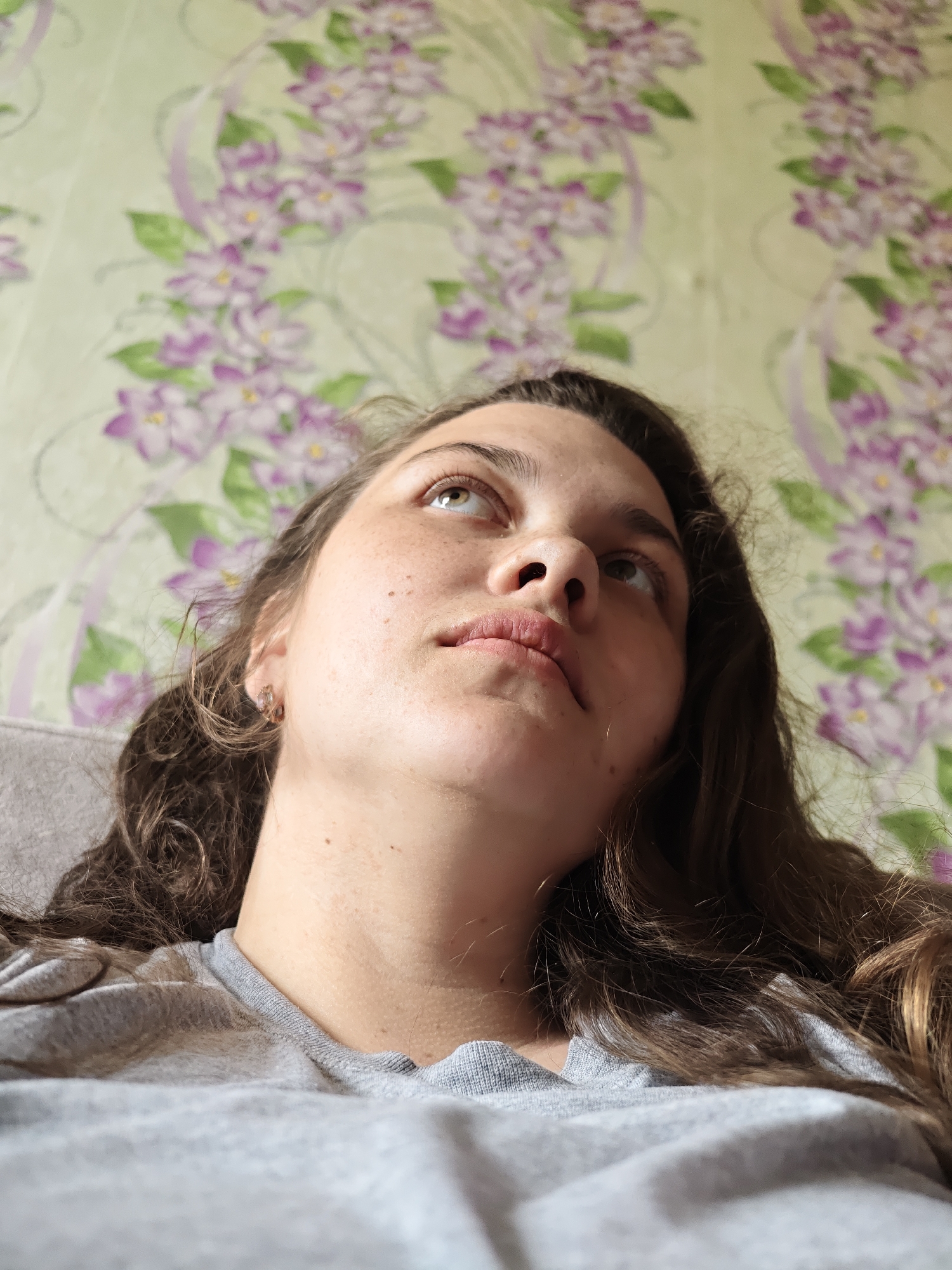 A low-angle, medium shot depicts a fair-skinned young woman with brown hair, looking upward. She has freckles on her face and is wearing a gray t-shirt. The background is a pale green wallpaper with a floral pattern of purple and white flowers. The photo was likely taken indoors. There is no evidence to suggest that the photo was taken in Уральское, Russia.