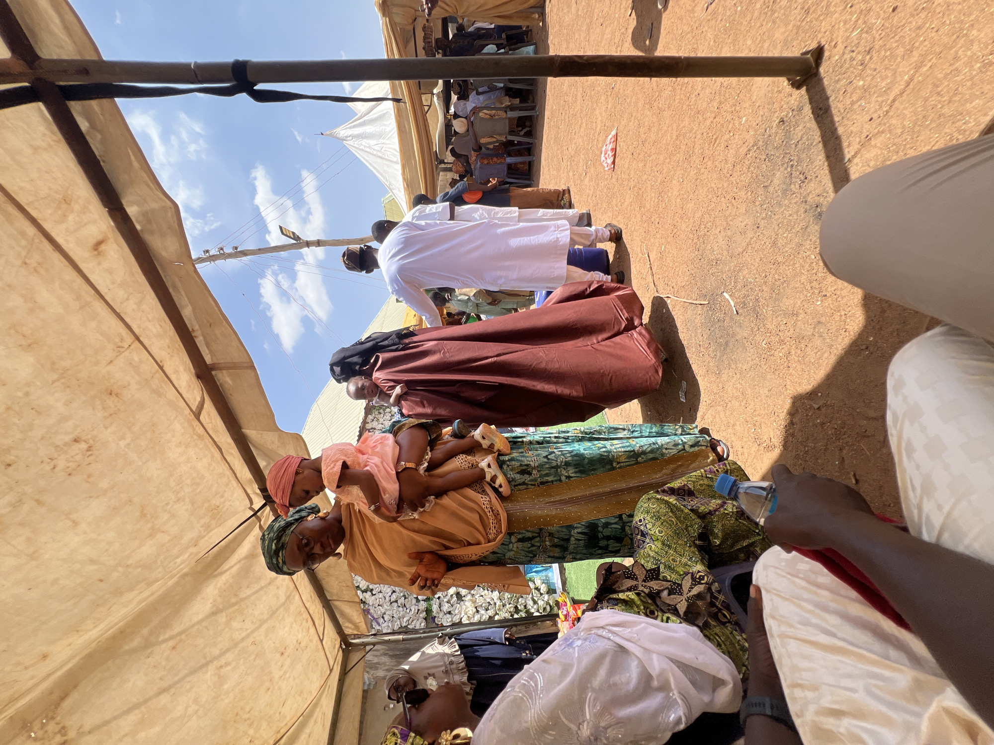 The image shows a gathering of people in Rigasa, Nigeria, likely attending an event under a tent. A woman wearing a colorful dress and headwrap is holding a child in her arms. Other individuals are visible in the background, including some wearing white robes and head coverings. The ground is dirt or sand, and additional tents are present. The atmosphere appears to be sunny.