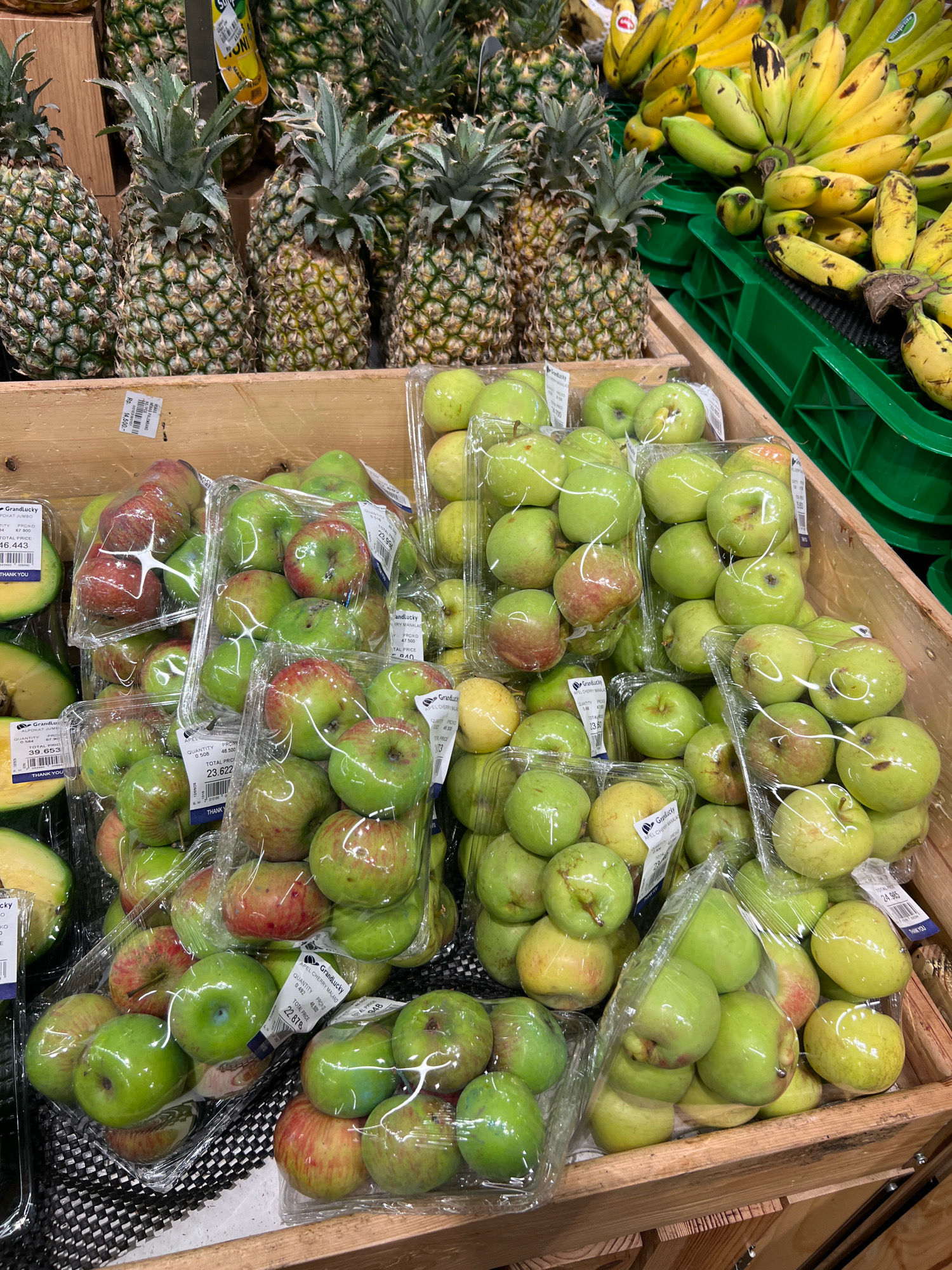 A close-up, eye-level shot captures a display of fresh fruit at a market in Kuta, Indonesia. In the foreground, plastic-wrapped containers of green and red apples are neatly arranged. Some apples are partially visible through the clear plastic packaging, showcasing their vibrant colors. Behind the apples, several pineapples with spiky green tops are stacked, their textured exteriors indicating ripeness. To the right, a large bunch of yellow bananas, some showing brown spots, spills out of a green plastic crate. The background is filled with more pineapples and bananas, creating a bountiful display of produce. The overall scene is well-lit, highlighting the freshness and variety of the fruits.