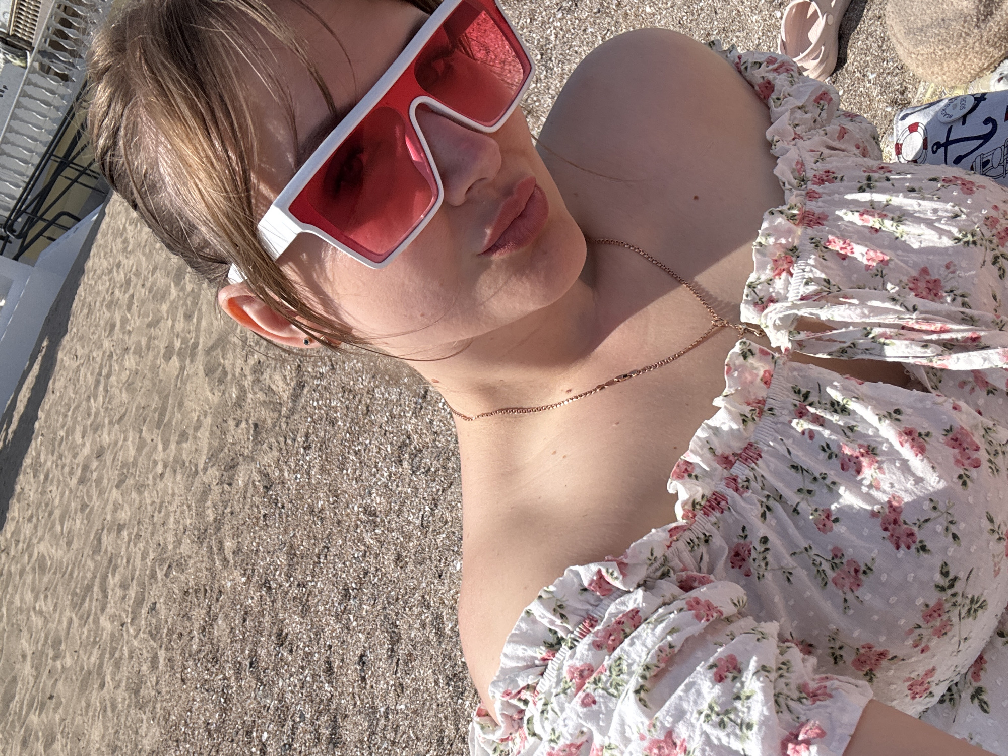 A woman wearing white sunglasses with red lenses and a floral top is lying on a sandy beach. She is facing toward the left, with her head tilted back. The sand is tan-colored. A necklace is visible around her neck. A light-colored beach bag and sandals are in the background, to the right. A white railing is in the upper-left corner. The scene appears to be outdoors, possibly on a beach in the city of Yevpatoriya, Ukraine.