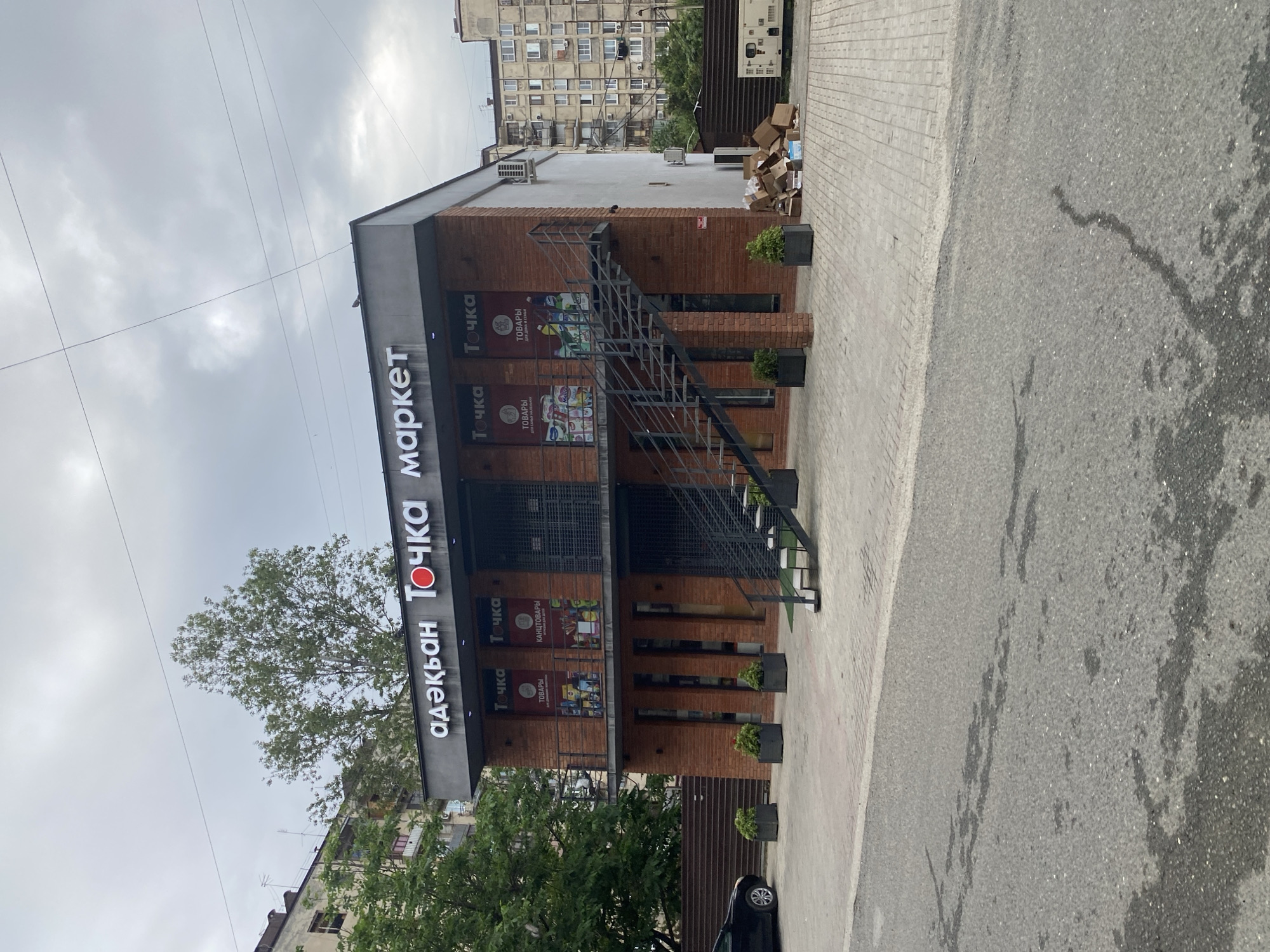 A street-level, exterior, full-shot photograph shows a modern, two-story brick building with a dark gray fascia and a large white sign that reads "аэкьян ТОЧКА маркет". The building features a metal staircase leading to a second-story balcony with a black metal railing. The ground floor has large windows with black frames, and the second floor has displays behind what appears to be rolled-down metal shutters. Two banners are visible on the second floor, one labeled "Точка" with the word "ТОВАРЫ" below it and "ДЛЯ ДОМА И СЕМЬИ" underneath that. Another banner on the second floor displays "Точка" with "КАНЦТОВАРЫ" and "Игрушки для детей" below. In front of the building, a paved area is visible with several planters containing small green bushes. To the left of the building, a black car is parked on the street. To the right of the building, a stack of cardboard boxes and some discarded material are piled next to a large white industrial unit. The sky is overcast with clouds. The building is situated in an urban environment, with residential apartment buildings visible in the background.