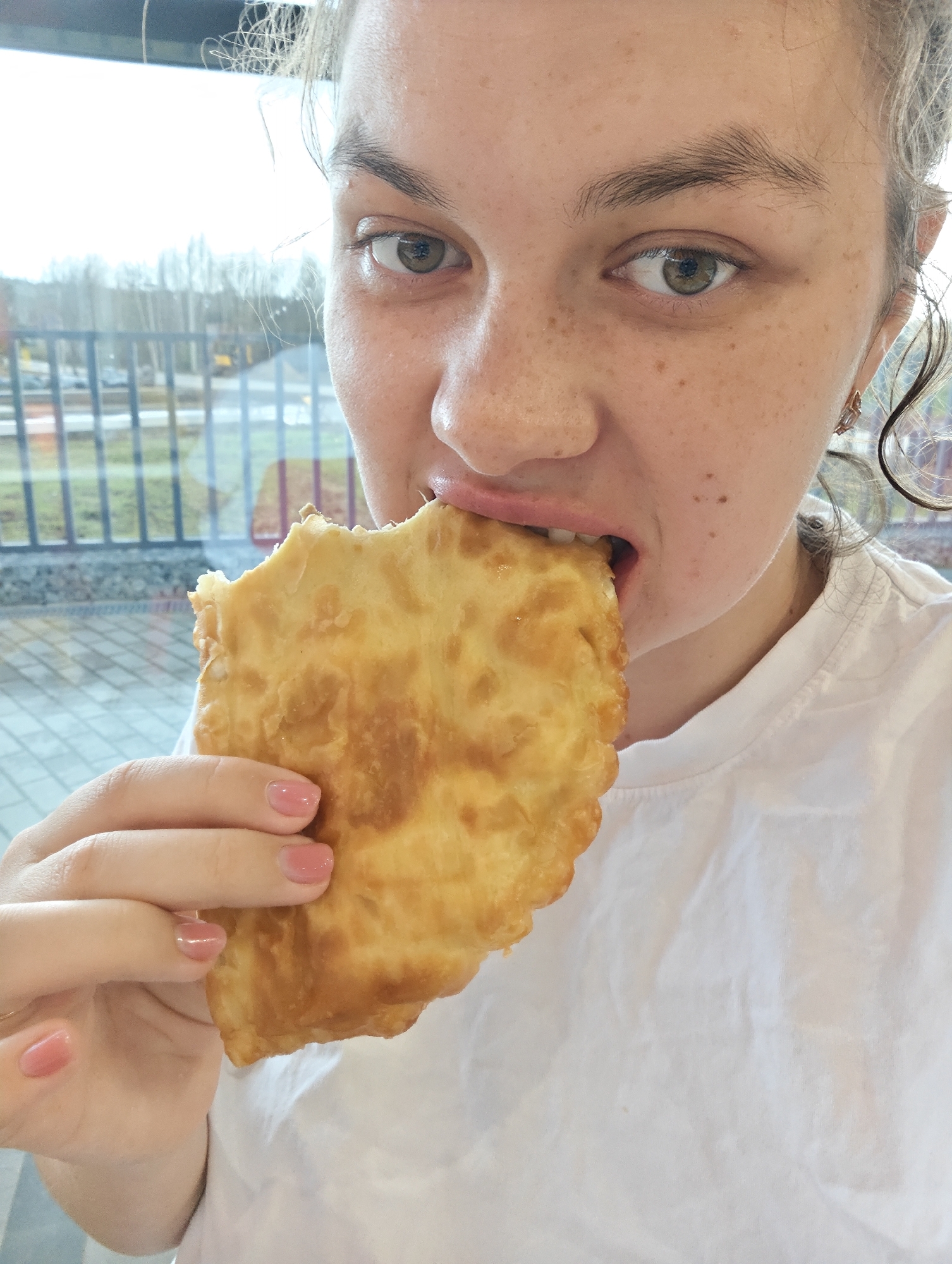 This close-up photograph captures a person in the midst of taking a bite from a large, golden-brown fried pastry, likely a cheburek. The subject, a young adult with light brown or hazel eyes, looks directly at the camera. Their face is adorned with visible freckles across the nose and cheeks. Light-colored, possibly curly hair frames their face, and they wear a simple white t-shirt. A small, intricate gold-toned earring is visible in one ear. The fingernails of the hand holding the pastry are painted with a soft pink polish.
The pastry itself is large, appearing flaky and crispy, suggesting it is freshly fried. The background consists of a large window looking out onto an outdoor scene. Beyond the window, bare trees suggest a cooler season like autumn or winter, alongside a paved area or road, and some indistinct distant structures. The lighting is bright but diffused, indicating an overcast daytime. The setting appears to be an indoor space, possibly a casual eatery or cafe, located in Votkinsk, Russia. No visible text is present in the image. The overall mood is one of focused enjoyment of the food.