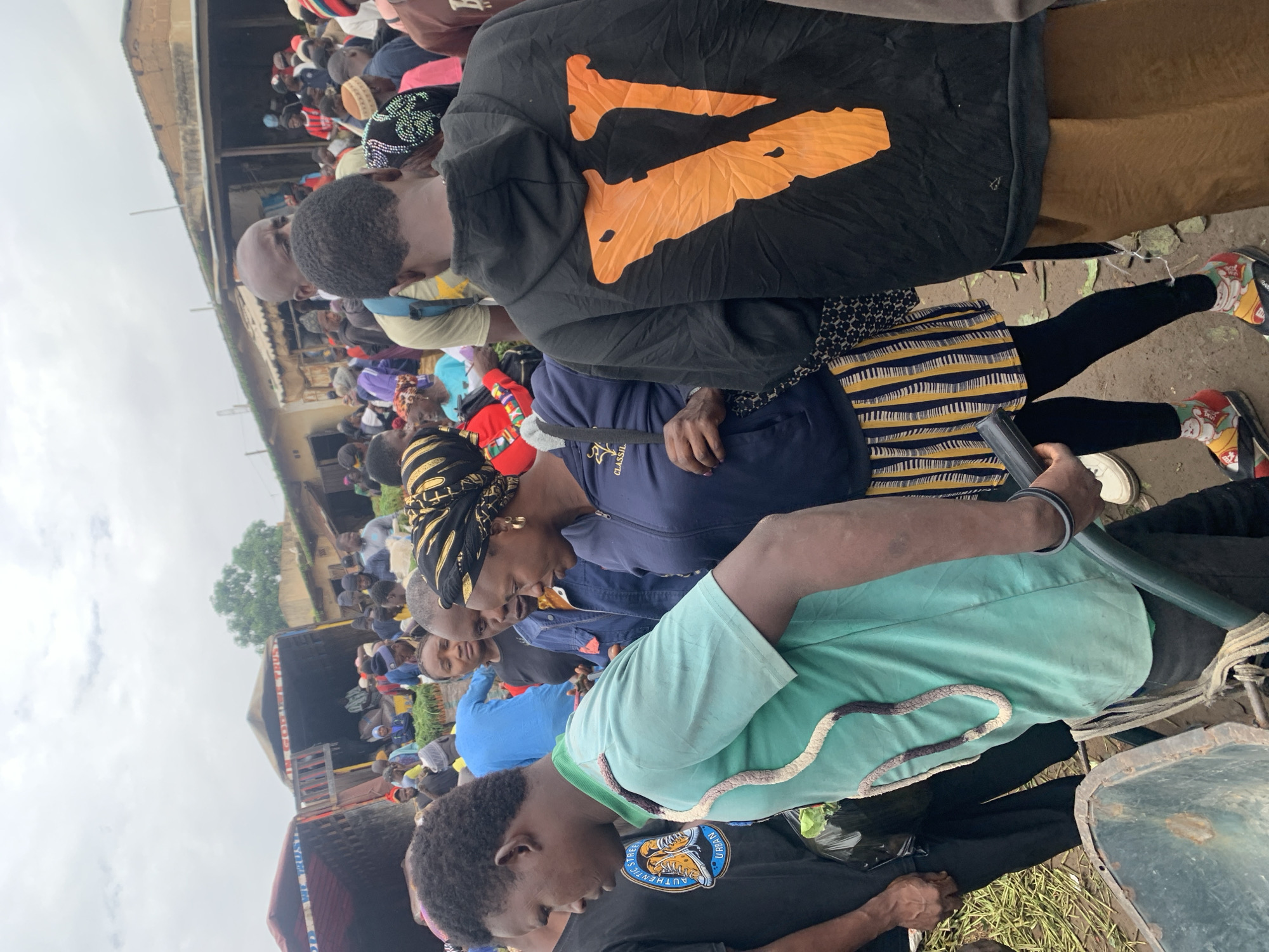 This media file depicts a busy outdoor market scene in Gura Top, Nigeria, likely during daylight hours with an overcast sky. The foreground is dominated by people interacting in a crowded environment, suggesting a bustling marketplace.
In the center, a person wearing a black hoodie with a large orange V-shaped design is visible. To their right, a person in a blue top and a patterned skirt, and another in a light blue t-shirt are prominent. The individuals are facing towards the right side of the frame, indicating a shared focus or activity. A person's arm in the foreground is holding a black object, possibly a phone or a tool.
The background reveals more people, stalls, and a truck, reinforcing the market setting. The presence of what appears to be produce or goods on the truck and the density of people suggest a commercial activity, likely buying and selling. The overall atmosphere appears active and communal. There is no visible text that can be deciphered. The weather appears to be cloudy, as indicated by the uniform grayness of the sky.