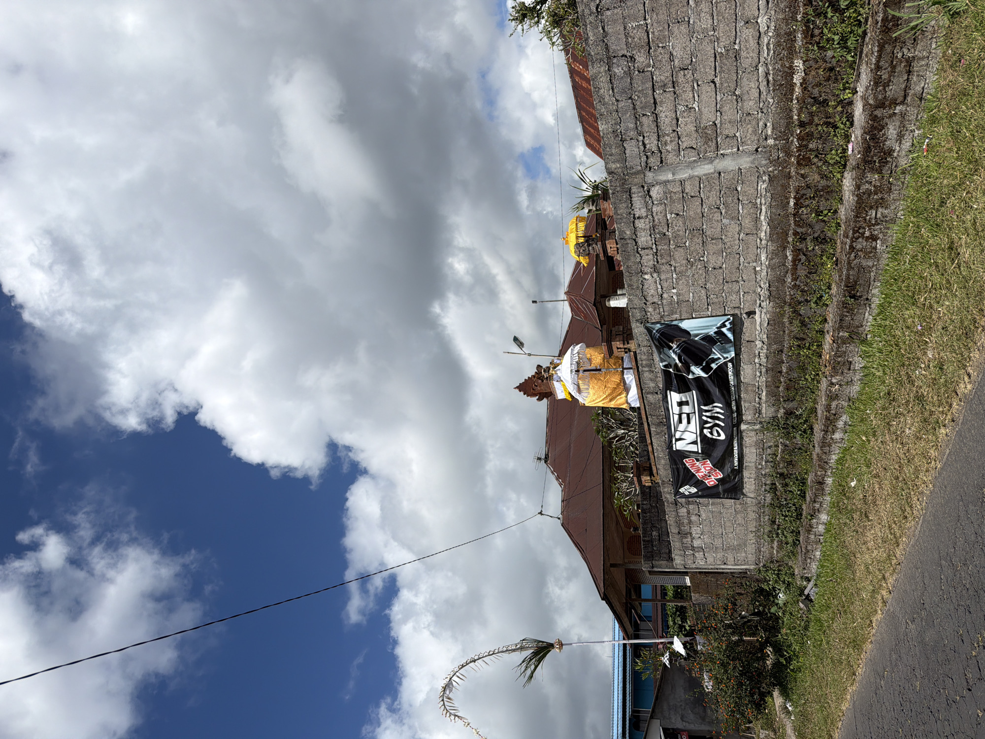 The image shows a street scene in Kintamani, Indonesia. In the foreground, a paved road runs along the left side of the frame, bordered by a strip of dry grass. A low, moss-covered stone wall runs parallel to the road, topped by a taller, more recently constructed cinder block wall. Attached to the cinder block wall is a black banner with white text that reads "NEO GYM". To the left of the banner, a narrow, leafy bush with red berries is visible.
Behind the wall, a building with a reddish-brown tiled roof is partially visible. A decorative Balinese umbrella, covered in yellow fabric, stands in front of the building, along with a tall, white ceremonial pole adorned with a green frond. The sky above is a bright blue, dotted with fluffy white clouds. The overall impression is of a somewhat rural or suburban area, with elements of local culture and commerce.