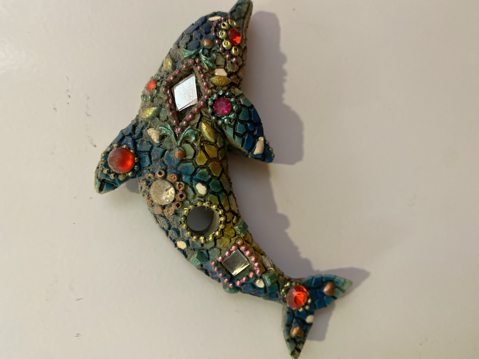 A mosaic dolphin ornament, adorned with colorful glass pieces and red, pink, and clear jewels, rests on a plain, light-colored surface. The dolphin is positioned horizontally, with its head facing to the right and its tail to the left. The surface on which it lies appears to be a wall, and a faint shadow of the dolphin is cast to its right. The ornament is the sole subject of the image.