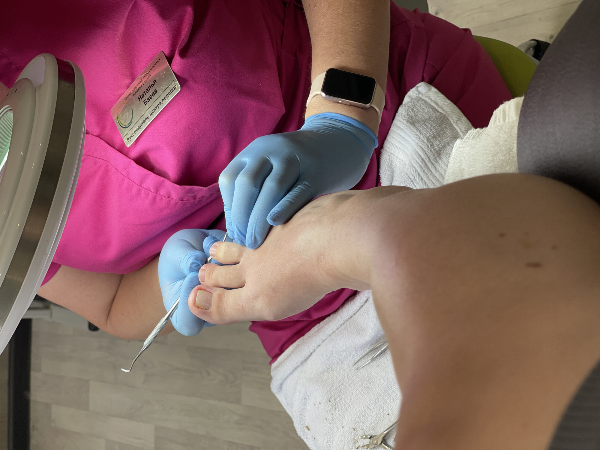 An overhead close-up shot shows a person wearing blue nitrile gloves performing a pedicure on a foot. The foot is resting on white towels, and various pedicure tools, including cuticle nippers and a metal file, are visible. The person is wearing a bright pink scrub top with a name tag that reads "Наталья Баева". A smartwatch with a light-colored band is worn on their wrist. The environment appears to be a salon, with a bright light fixture overhead and a blurred background of a tiled floor.