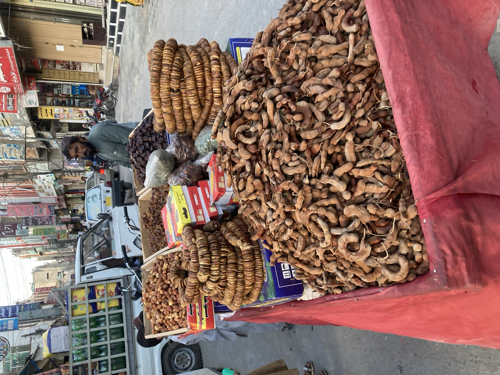 The image depicts a street vendor in Quetta City Tehsil, Pakistan. The vendor, a bearded man wearing a skullcap and green shirt, stands behind a table laden with dried fruits and nuts. The table is covered with a red cloth and displays large piles of tamarind pods, dried figs strung into rings, and boxes of dates.
The scene is set on a bustling street, with storefronts and signage visible in the background. A white delivery vehicle is parked nearby, with shelves containing various food and beverage products. Other vehicles and pedestrians can be seen further down the street, suggesting a busy commercial area. The overall impression is of a typical marketplace scene in an urban setting in Pakistan.